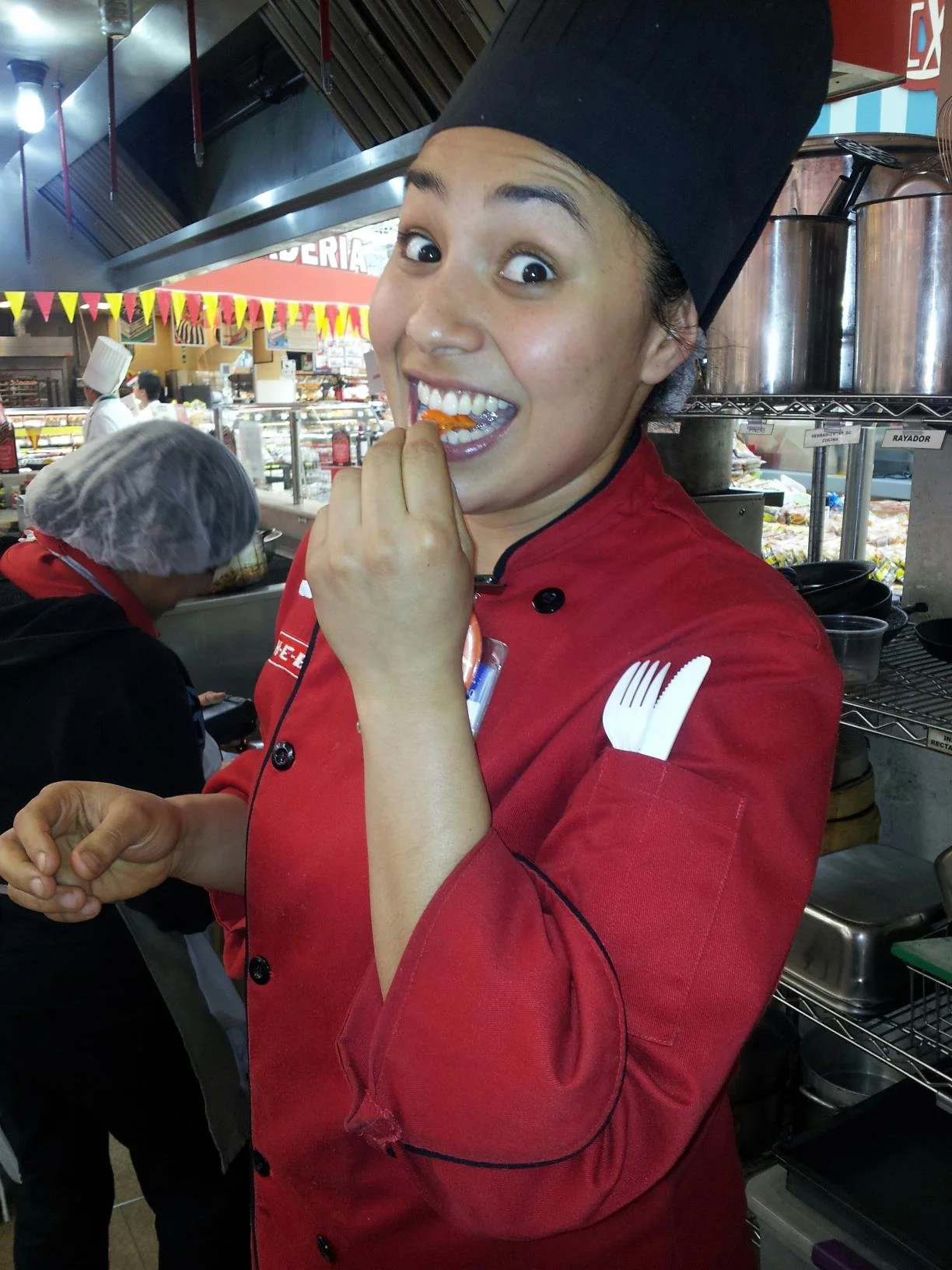 A young Chef Fernanda Reyes in a red HEB expo chef uniform and black hat is smiling and biting into a hot chili pepper, with a busy kitchen in the background.