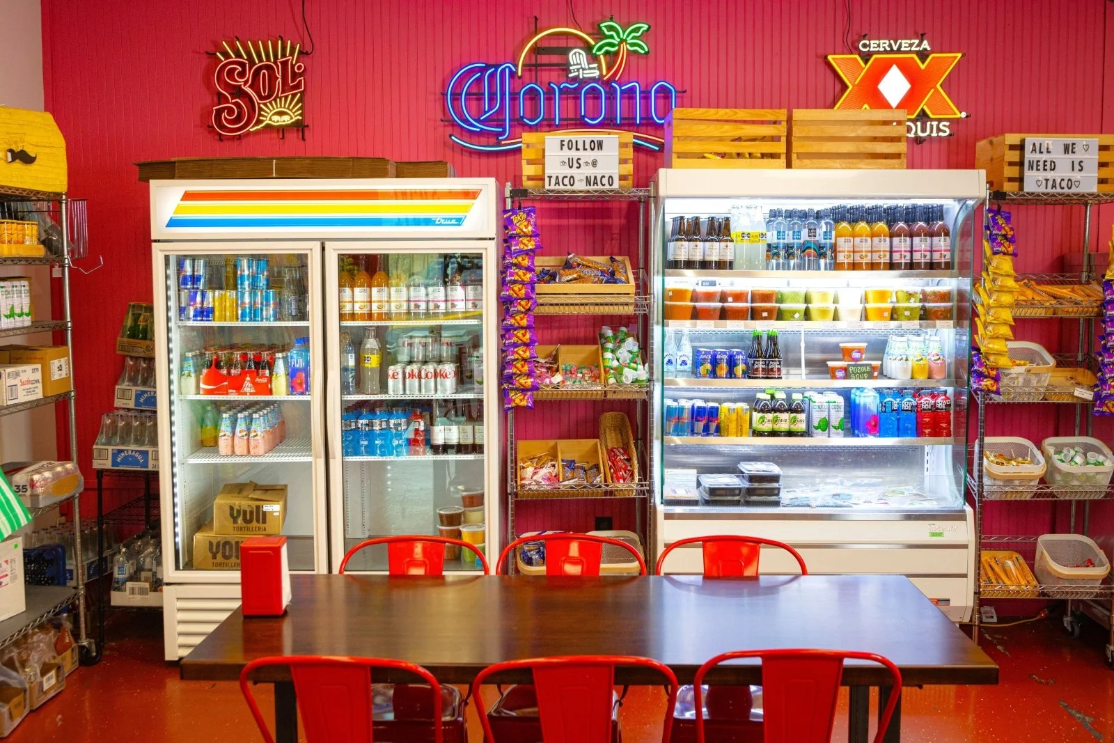A small convenience store with a table and six red chairs in front. Behind the table, there are two freezers stocked with various drinks, snack shelves, and a neon sign that says 'Corona' on a pink wall with decorative signs for 'Sol' beer and 'XX' beer. There are shelves on either side with snacks and other products.