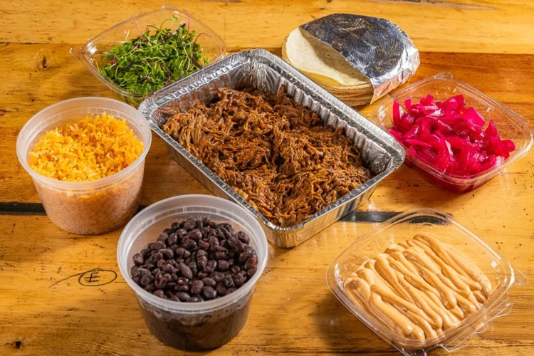 Shredded beef in a foil tray, black beans, shredded cheese, pickled red onions, shredded lettuce, sour cream, a small container of cotton candy, and a bread roll on a wooden table.