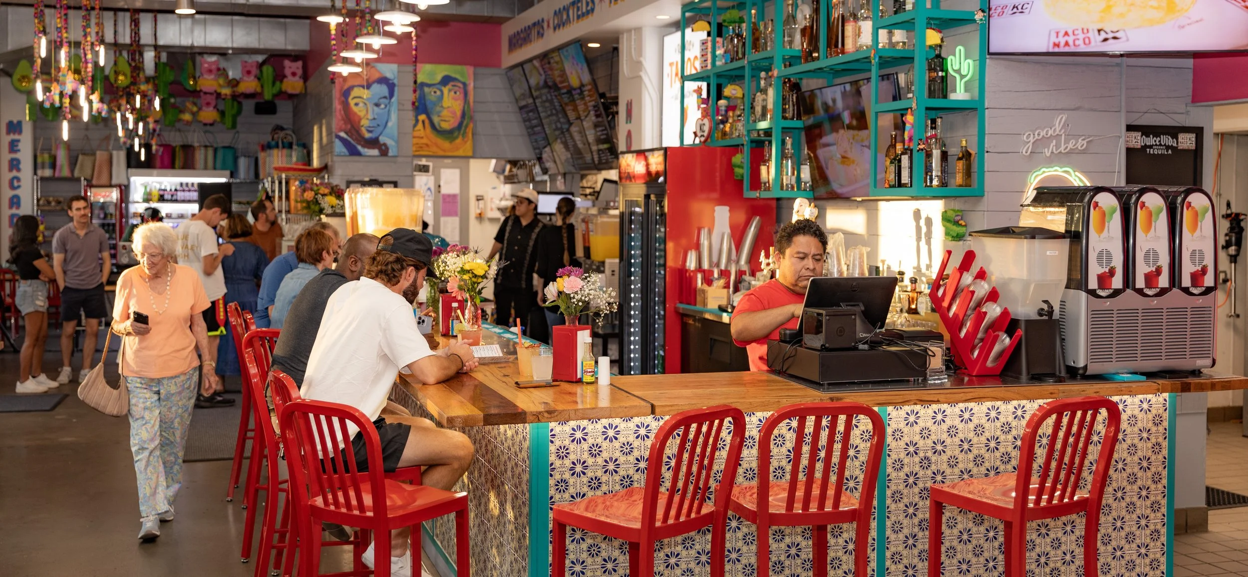 Colorful Mexican restaurant interior with diners, red chairs, tiled counter, tequila shelves, and frozen drink machines
