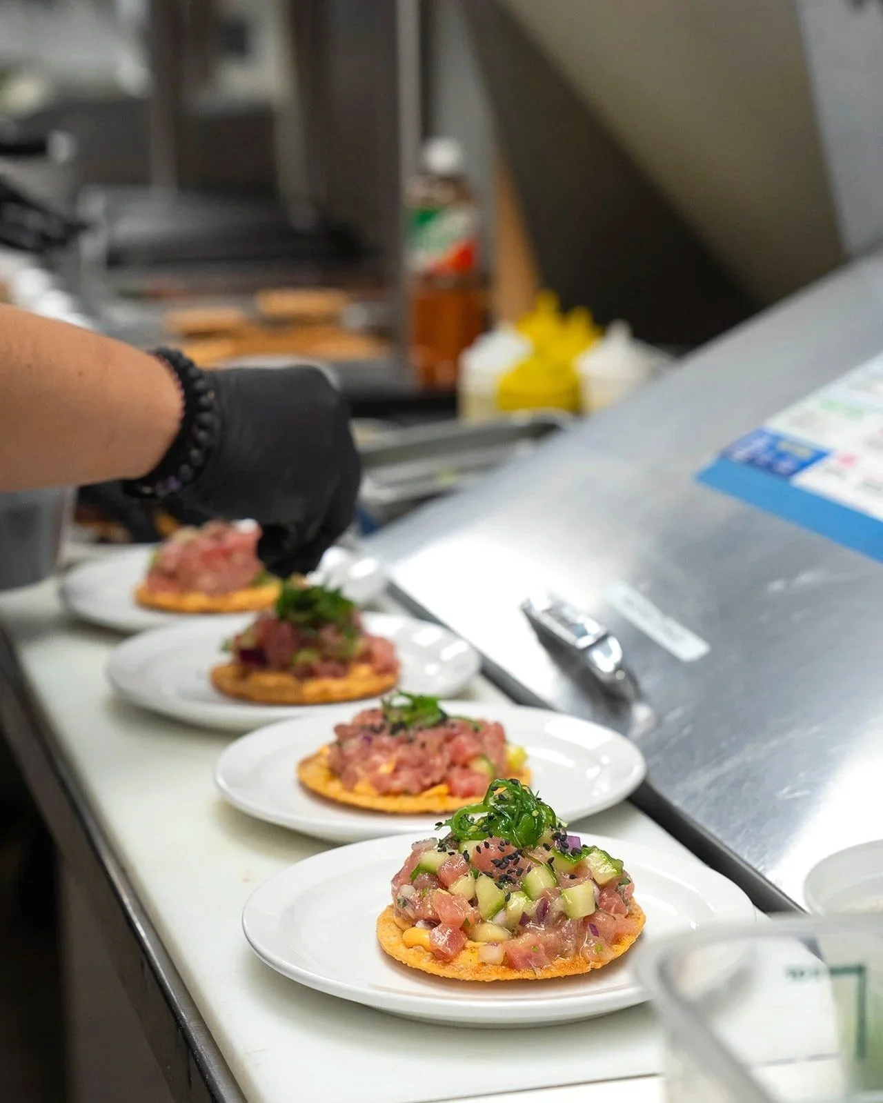 A row of plates with tuna tartare topped with green onions and black sesame seeds on a stainless steel countertop in a kitchen.