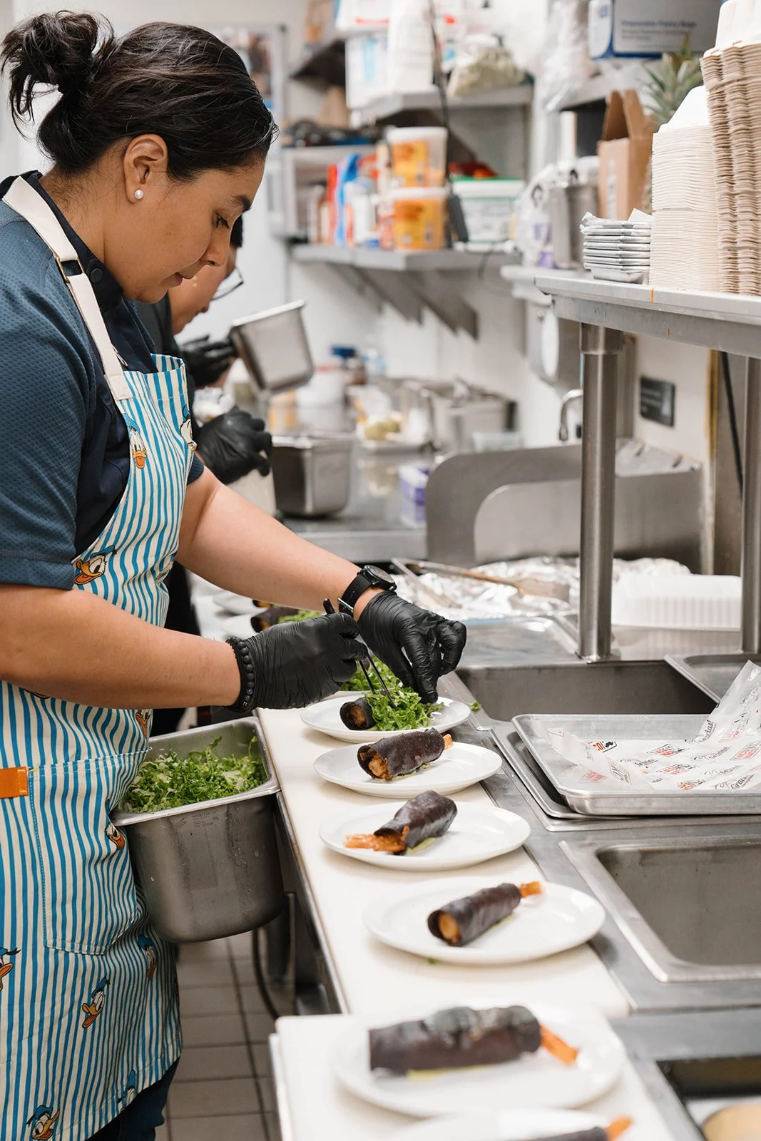 A woman in a striped apron and black gloves prepares chef-forward dishes in a commercial kitchen, garnishing the plated dished with greens