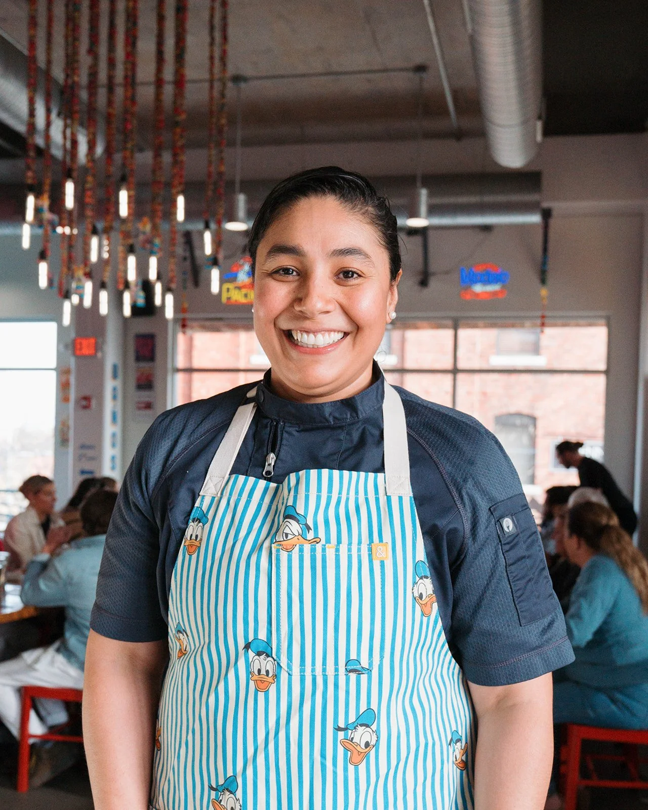 A smiling woman wearing a Donald Duck apron in a restaurant with diners in the background.
