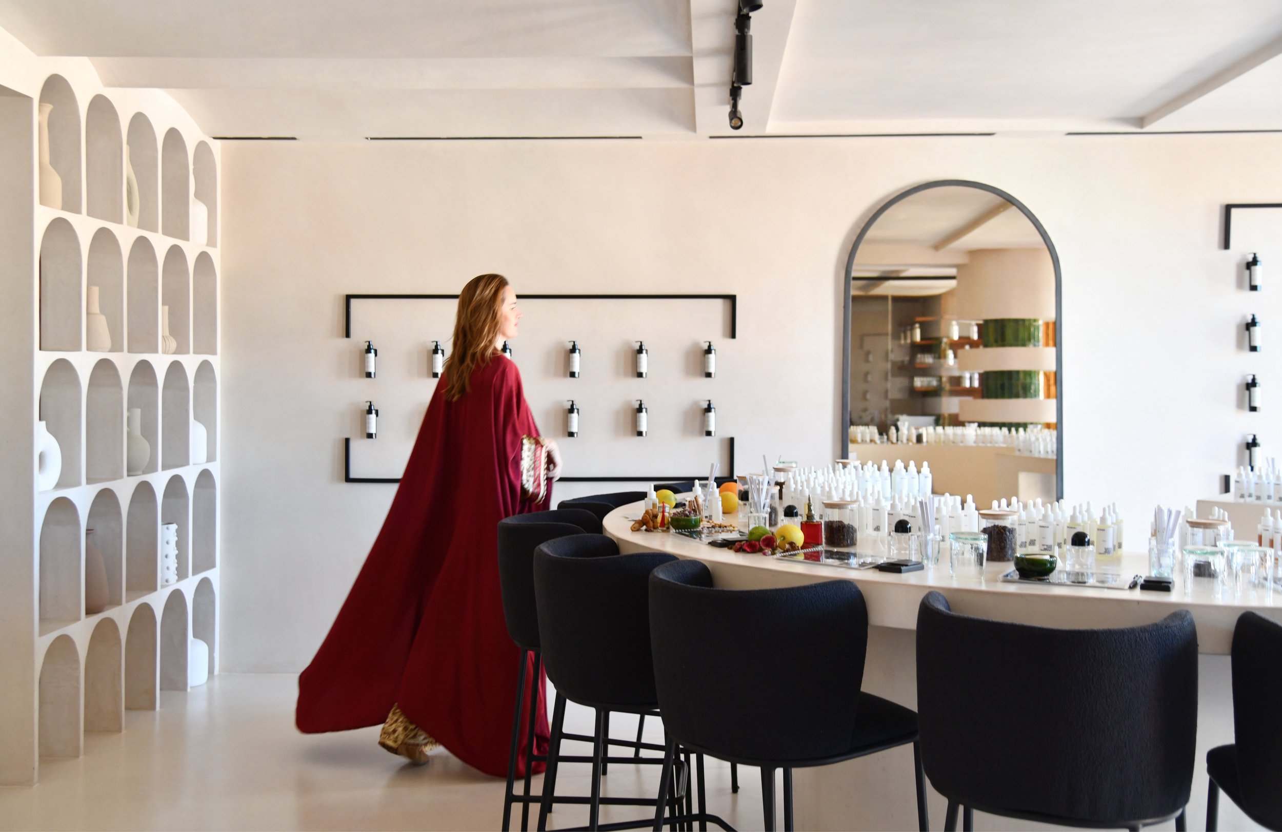 A woman in a red flowing dress walking in a modern, minimalist interior space with a display of bottles on the wall and a long table with various items, including fruits and bottles.
