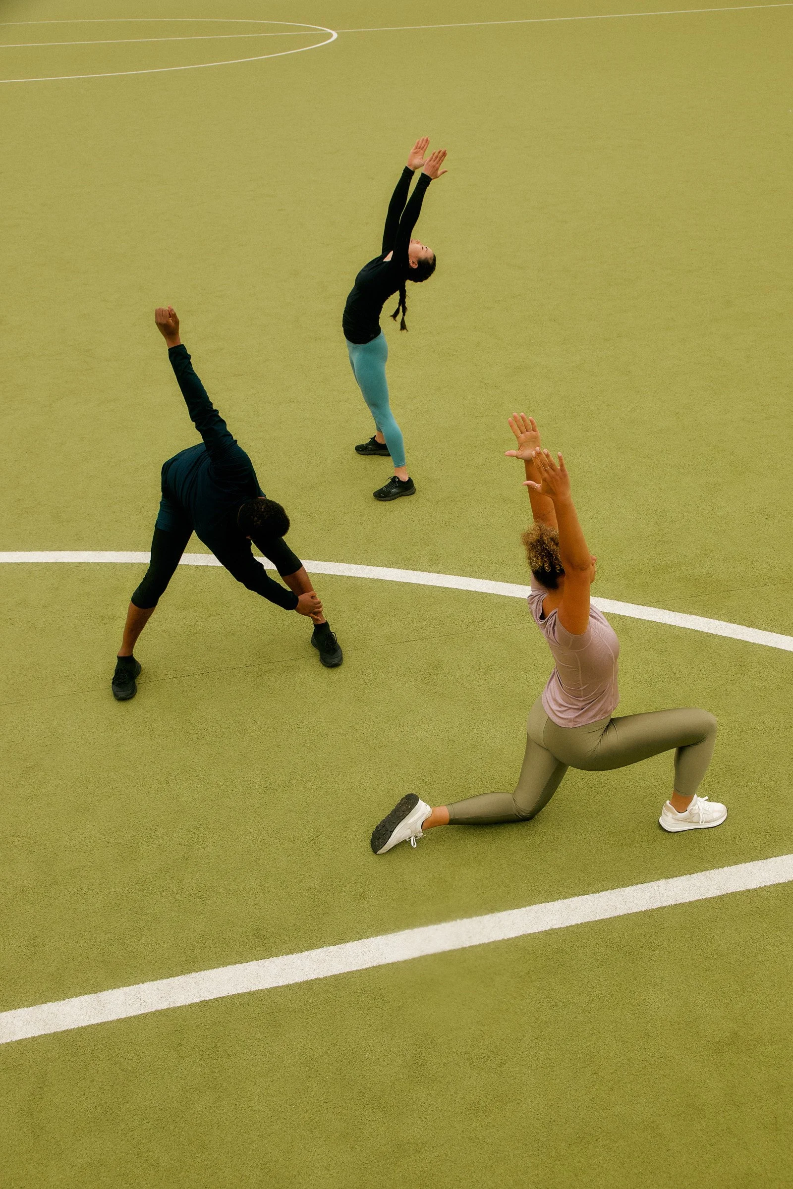 Group of women practicing yoga outdoors on grass in the morning, seated in a cross-legged position with their eyes closed, wearing comfortable exercise clothing with mountains and trees in the background.