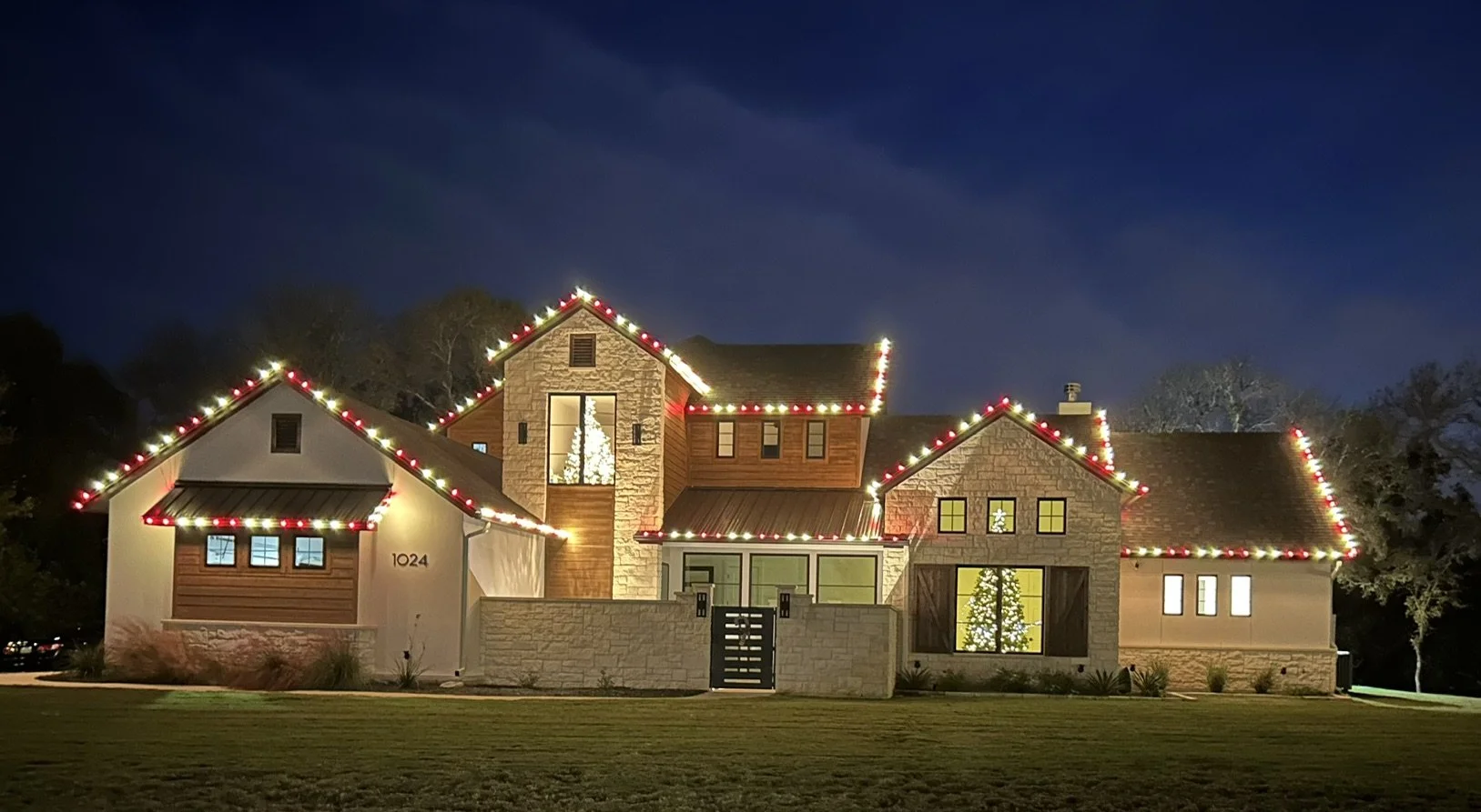 A large house decorated with Christmas lights on the roofline and a lit Christmas tree inside visible through the window at night.