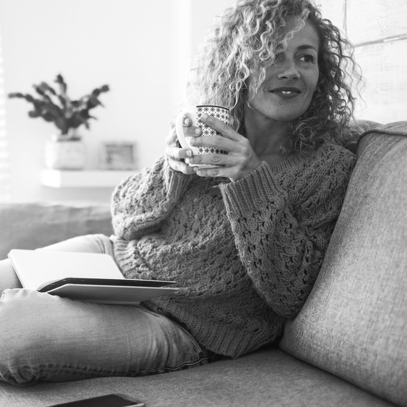 A woman with curly hair sitting on a couch, holding a patterned mug, and smiling. She has an open notebook on her lap, and there is a blurred background with a potted plant.