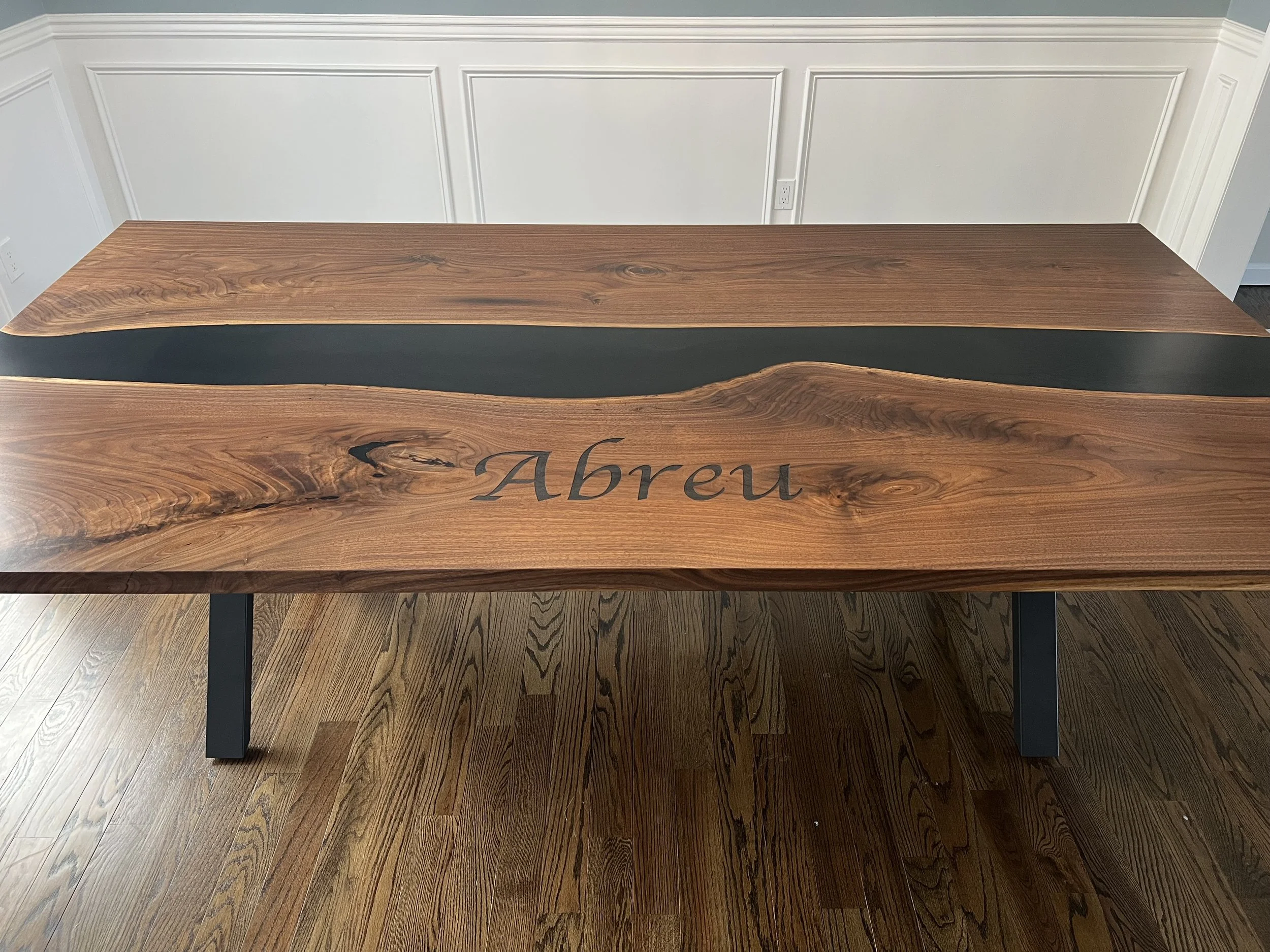Wooden table with a black resin inlay and the name 'Abreu' engraved on it, set on a hardwood floor with white paneled walls in the background.
