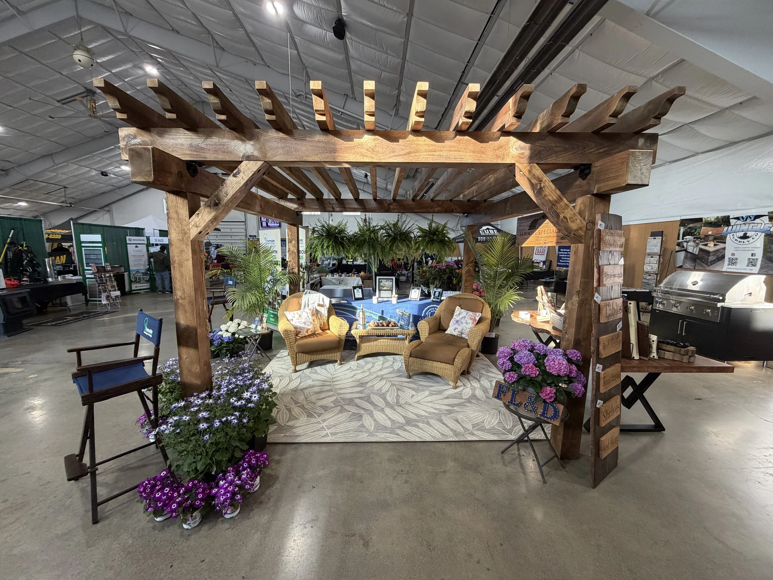 Indoor event space with a wooden pergola structure, two wicker armchairs with pillows, plants, and decorative flowers, set up on a patterned rug.