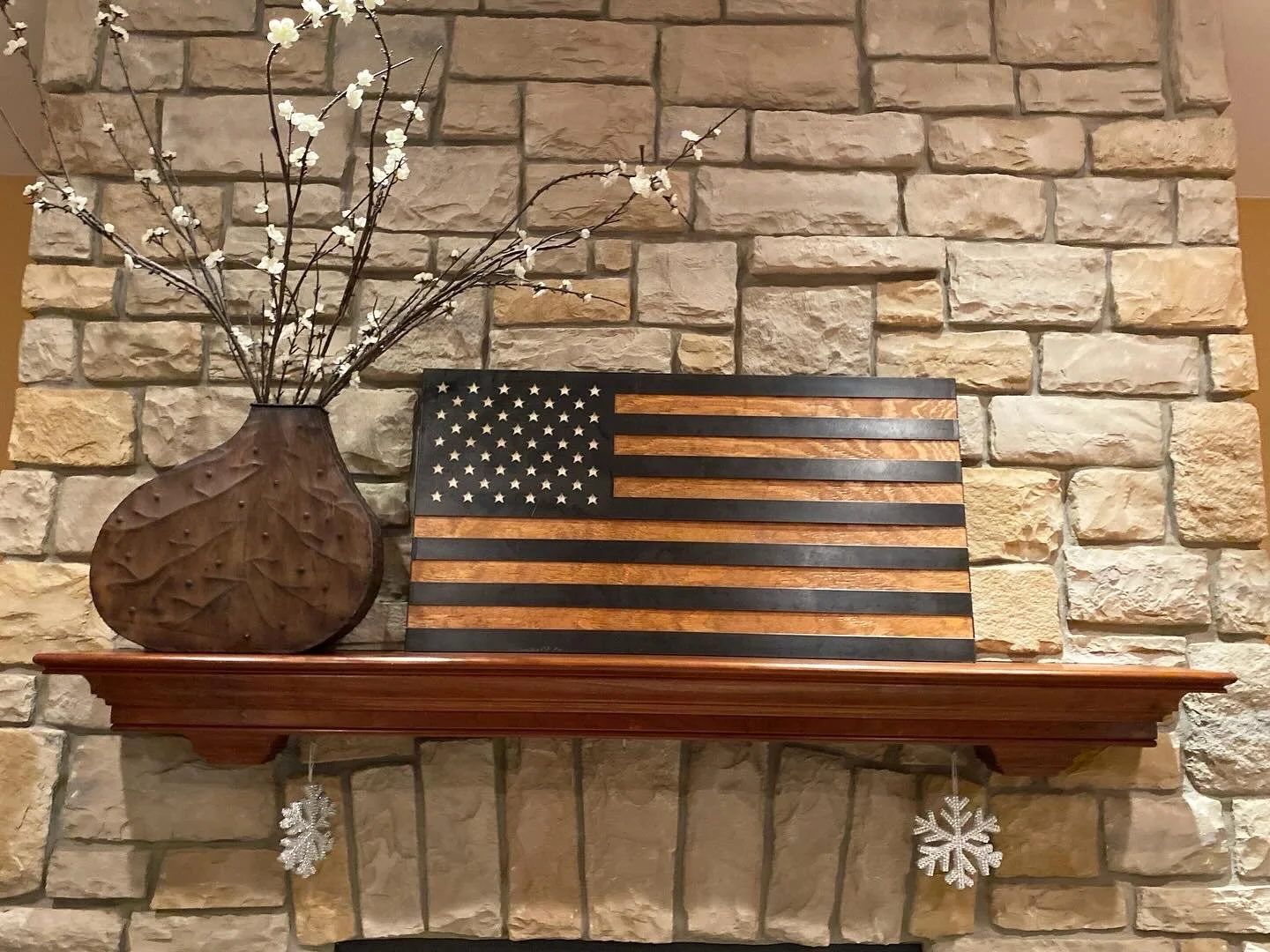 A wooden shelf on a stone wall holds a brown vase with white flowers and a wooden American flag decorative piece. Silver snowflake ornaments hang from the shelf.