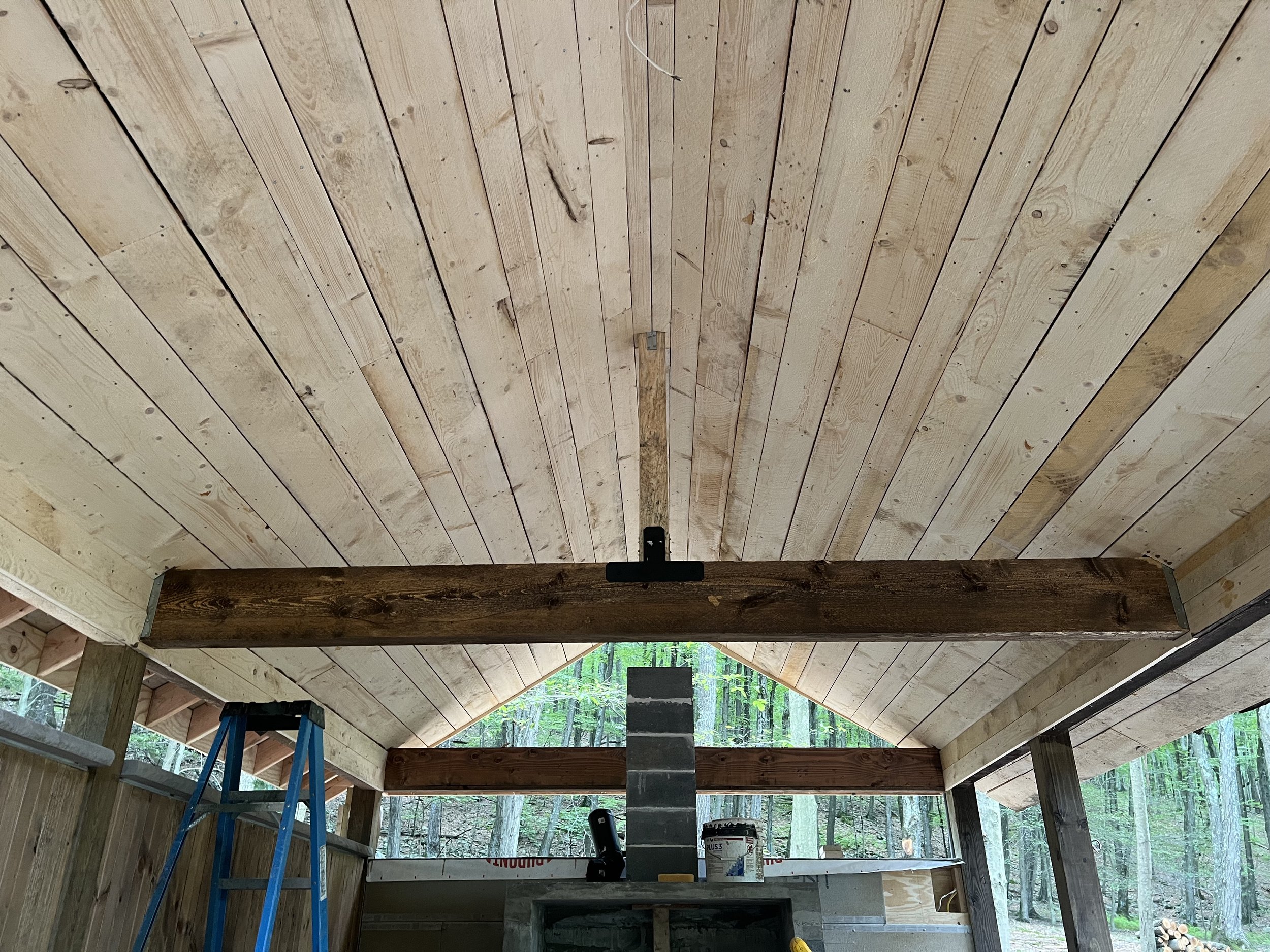 Interior of a wooden structure under construction, showing a vaulted ceiling made of light-colored wooden planks and dark wooden beams. A blue ladder is leaning against the wall, and construction tools and materials are visible on a counter below.