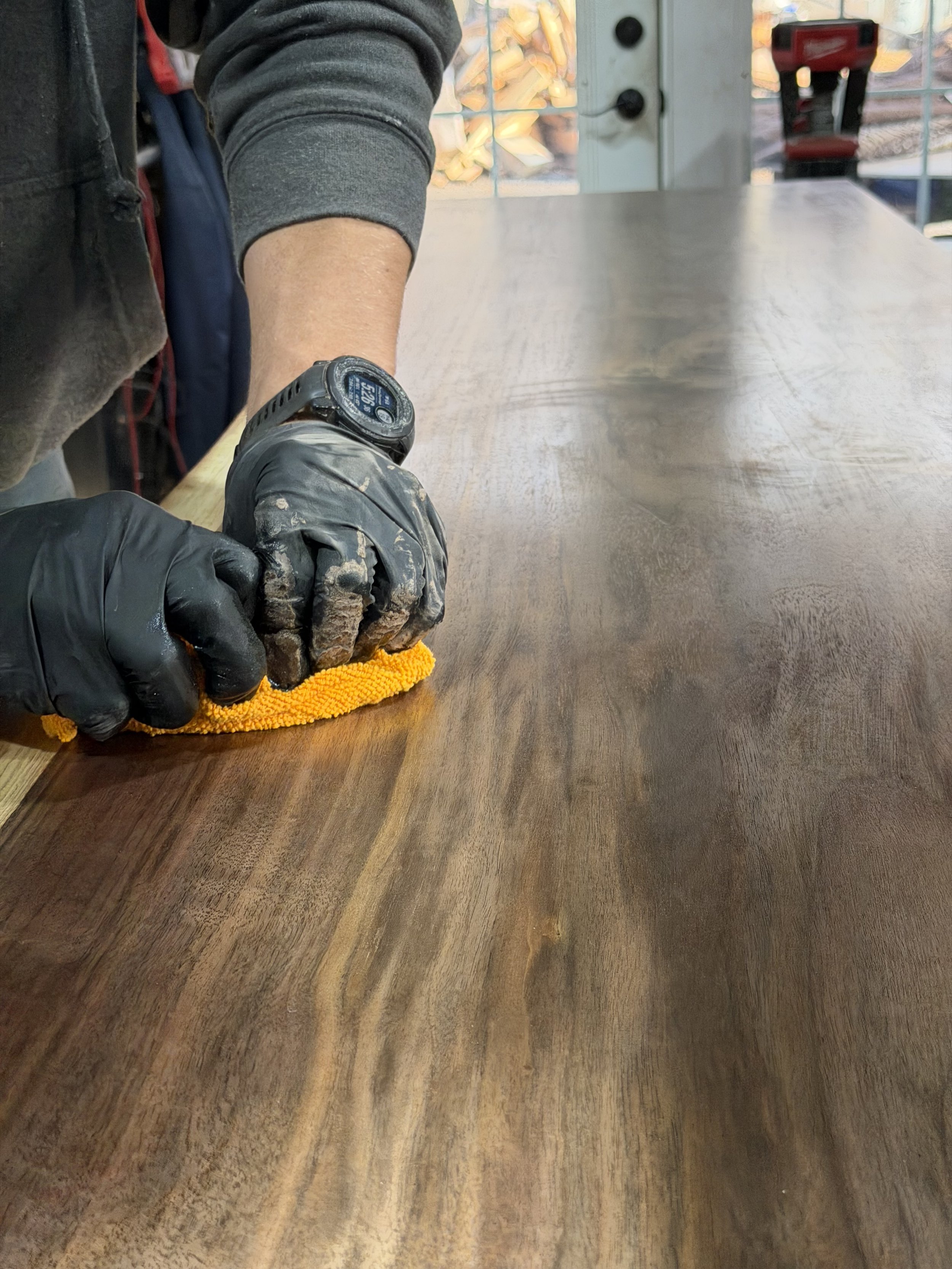 Person sanding or polishing a wooden surface with a cloth while wearing black gloves and a watch.