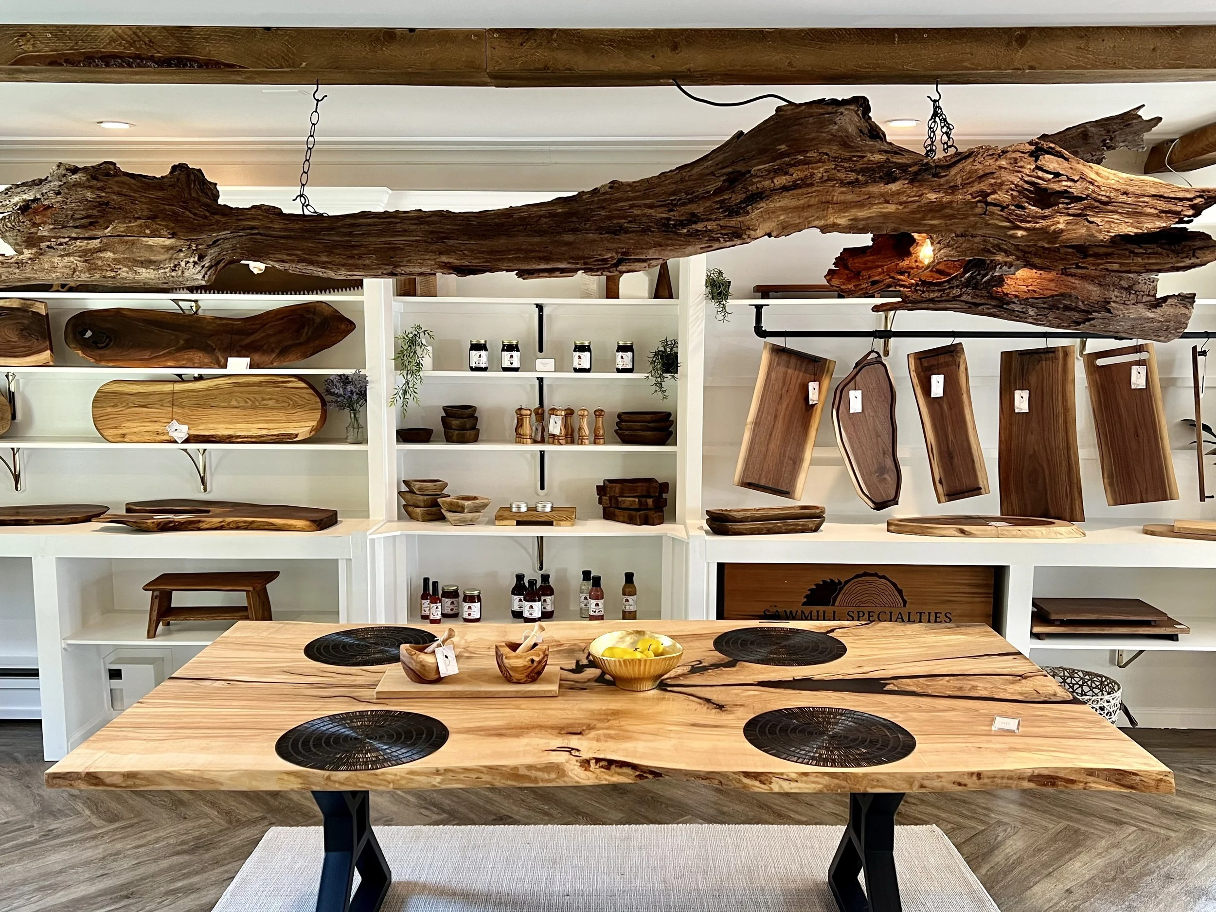 Wooden furniture store display with slices of wood, cutting boards, bowls, and a table with a natural edge, arranged on white shelves and hanging on the wall.