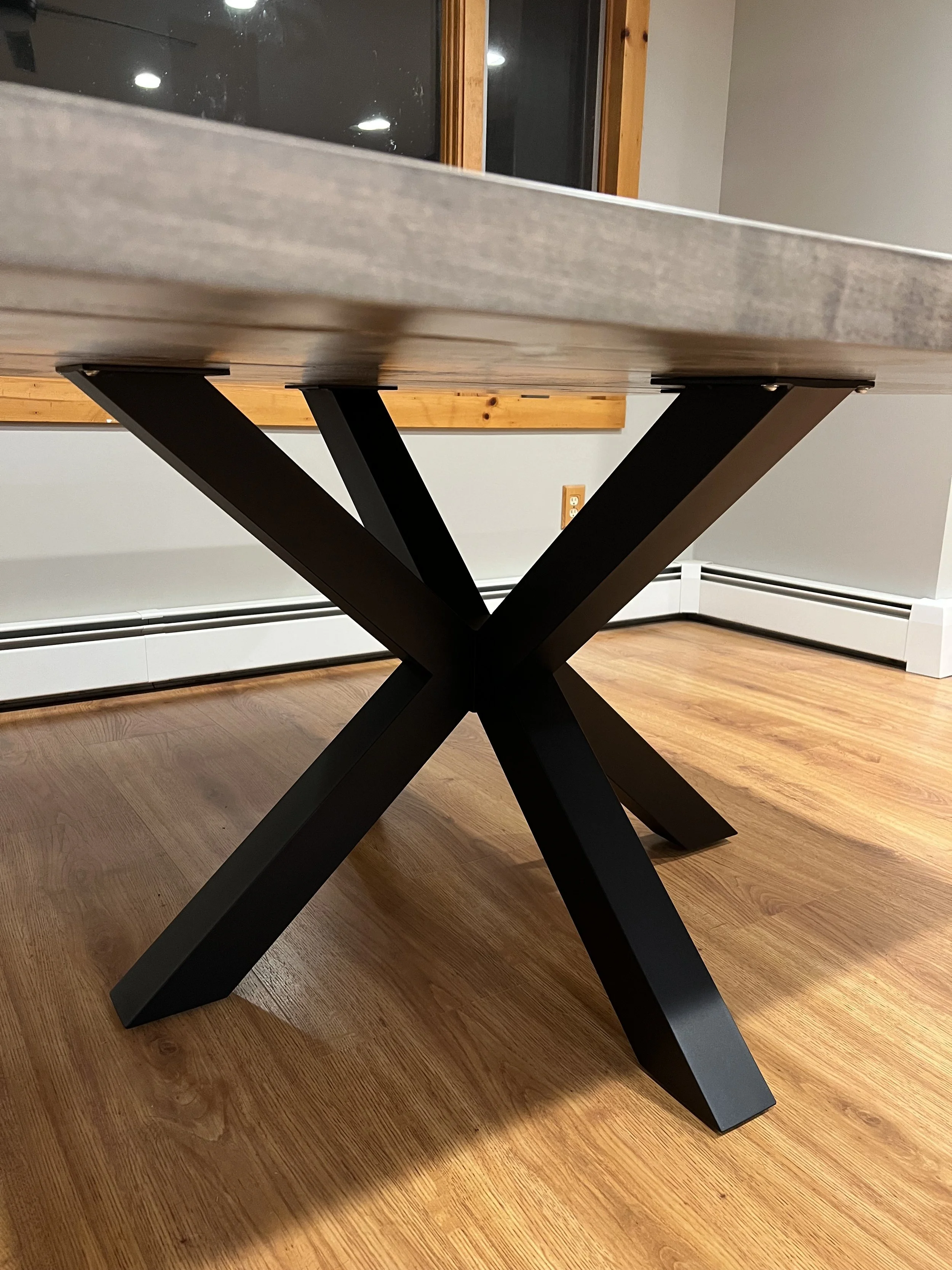 Close-up of a modern table with a wooden top and black metal X-shaped legs in a room with wooden flooring and a wall with power outlets.