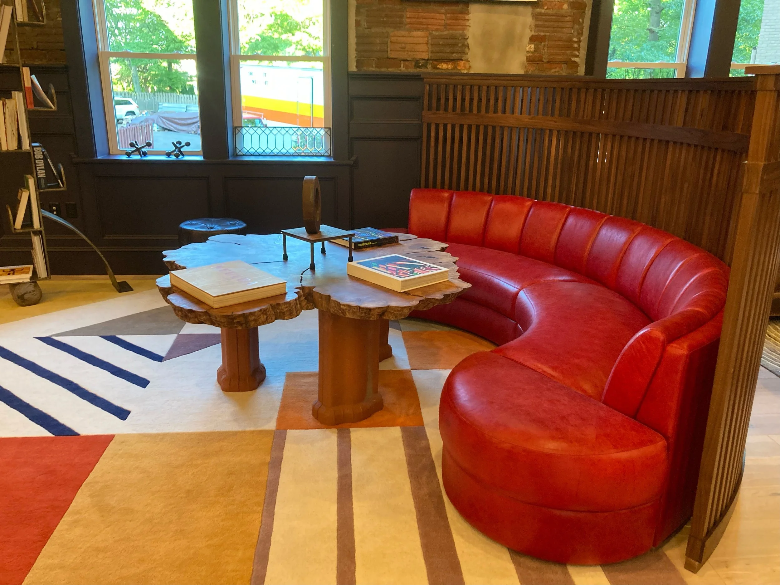 A cozy reading nook with a curved red leather sofa, a rustic wooden coffee table with books and decorative items, a multicolored geometric rug, large windows with a view of outdoor greenery, dark wood wall paneling, and bookshelves nearby.