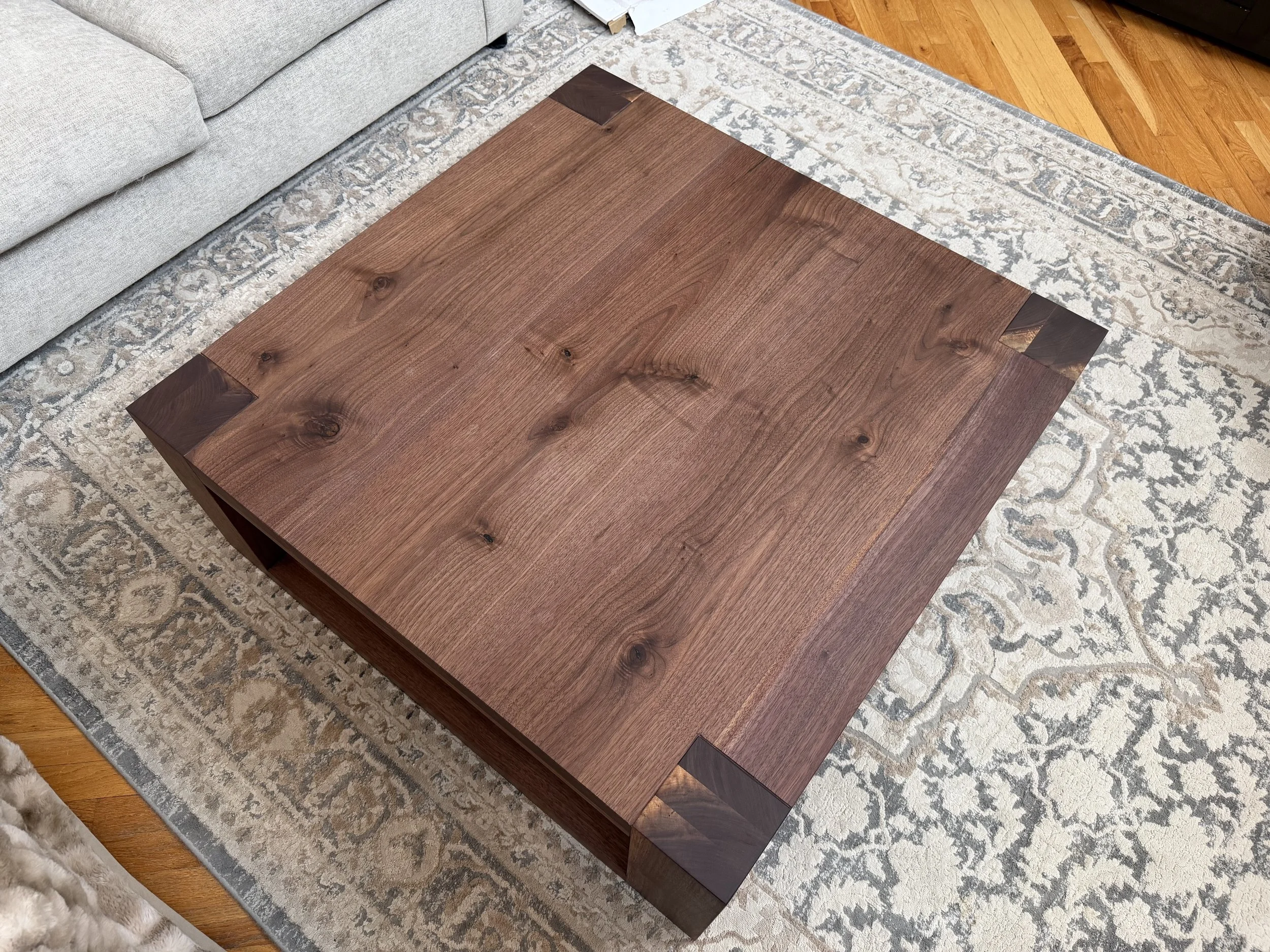 Wooden coffee table with a square top and dark corner accents, placed on a patterned area rug in a living room.