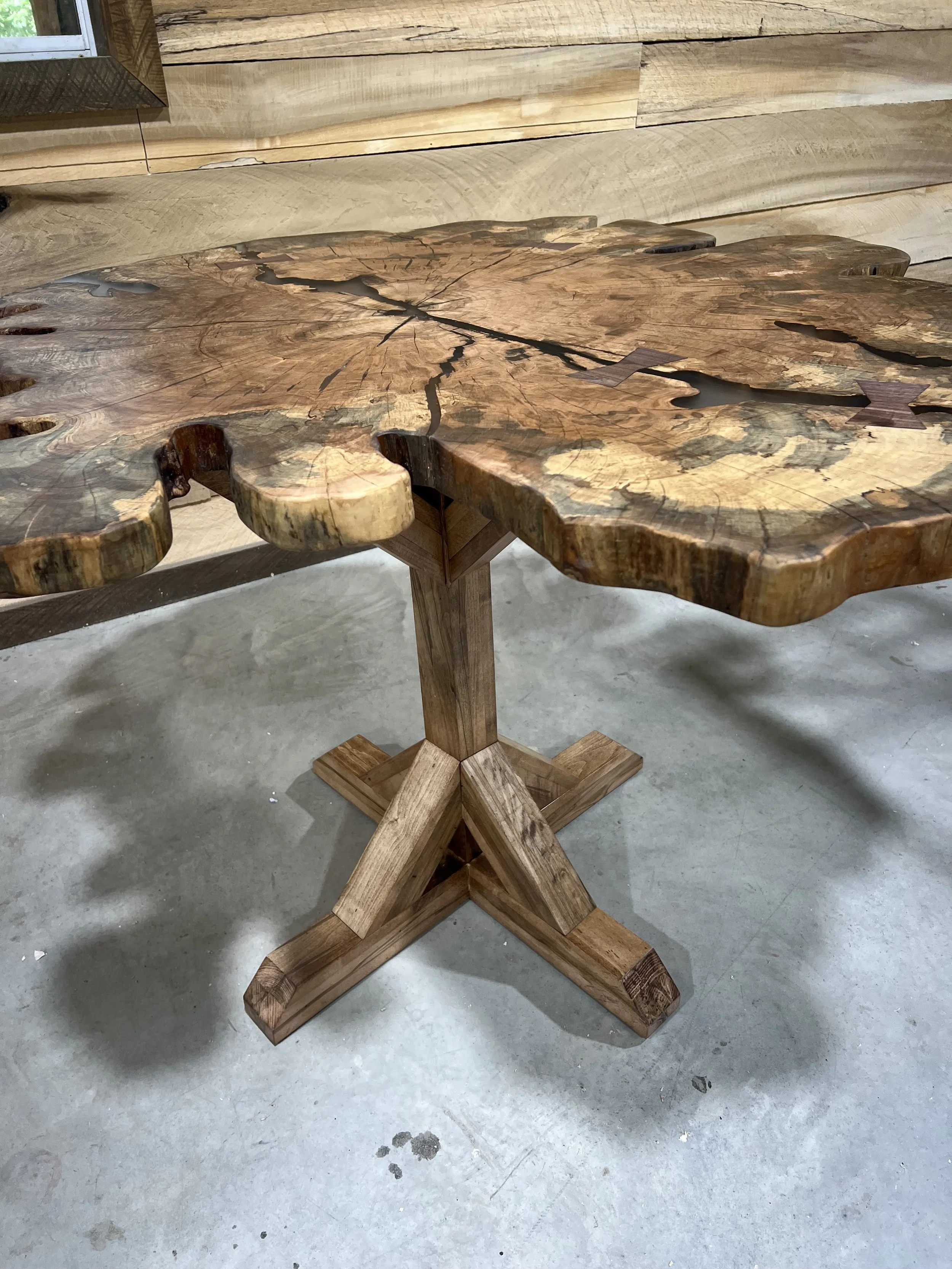 A wooden table made from a large, natural, live-edge slab with visible cracks and growth rings, supported by a sturdy, cross-legged wooden base, placed on a concrete floor.