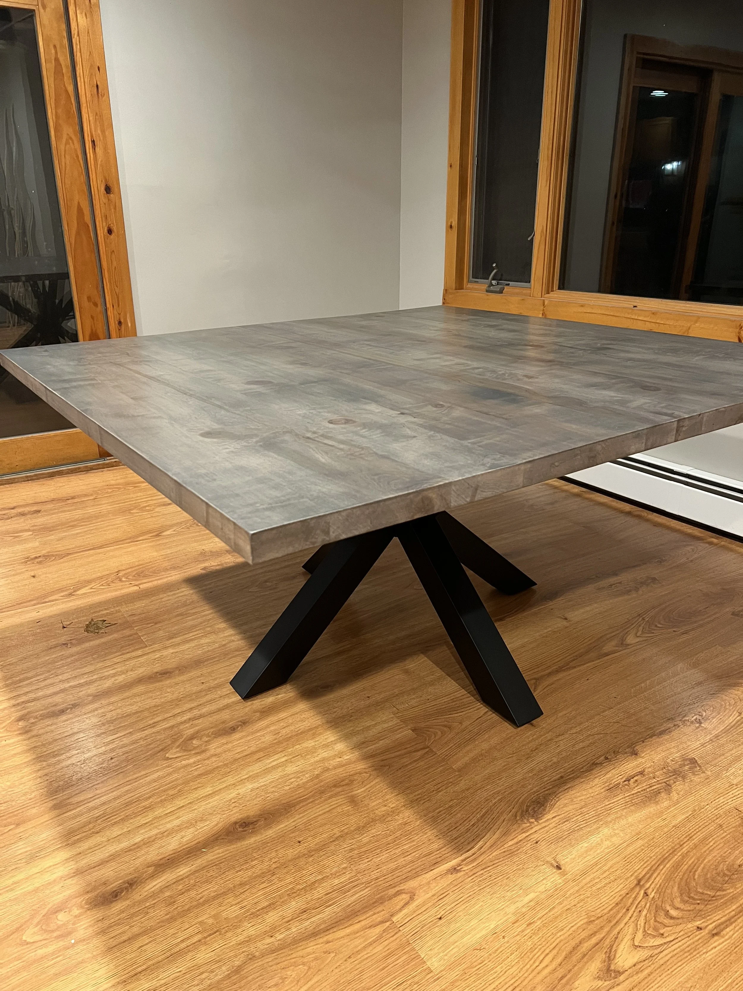 A wooden table with a grayish wood top and black angled legs in a room with wooden flooring, next to windows with wooden frames.
