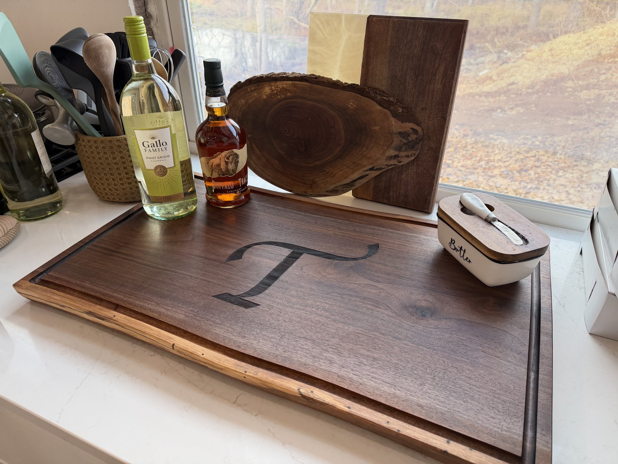 Wooden serving board with a monogrammed letter 'T' logo, a butter dish with a spoon, an artisan cutting board, bottles of wine and liquor, and kitchen utensils near a window.
