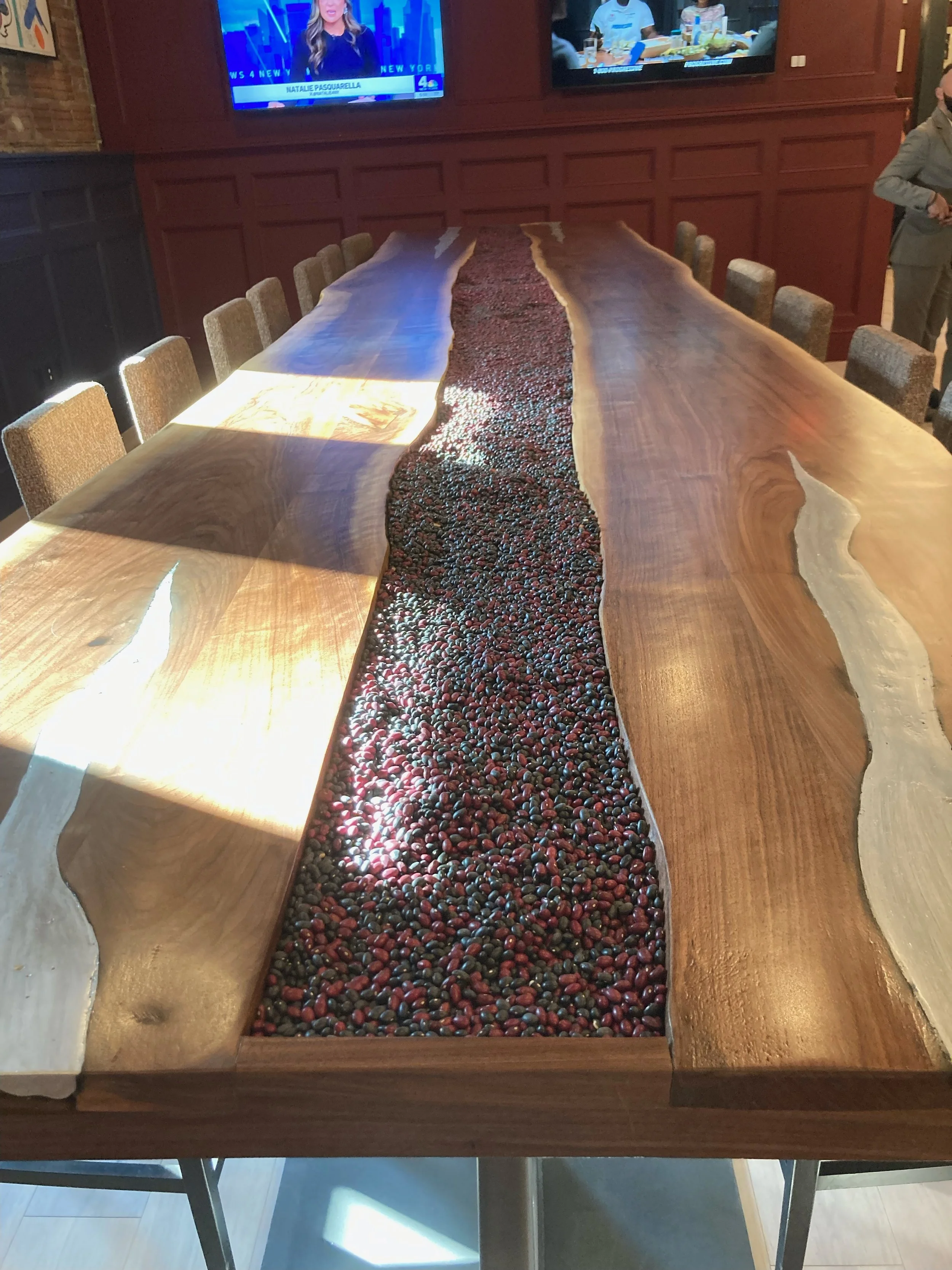 A large wooden conference table with a central inlay of colorful beans or seeds, surrounded by chairs in a restaurant or meeting room with two television screens on the wall.