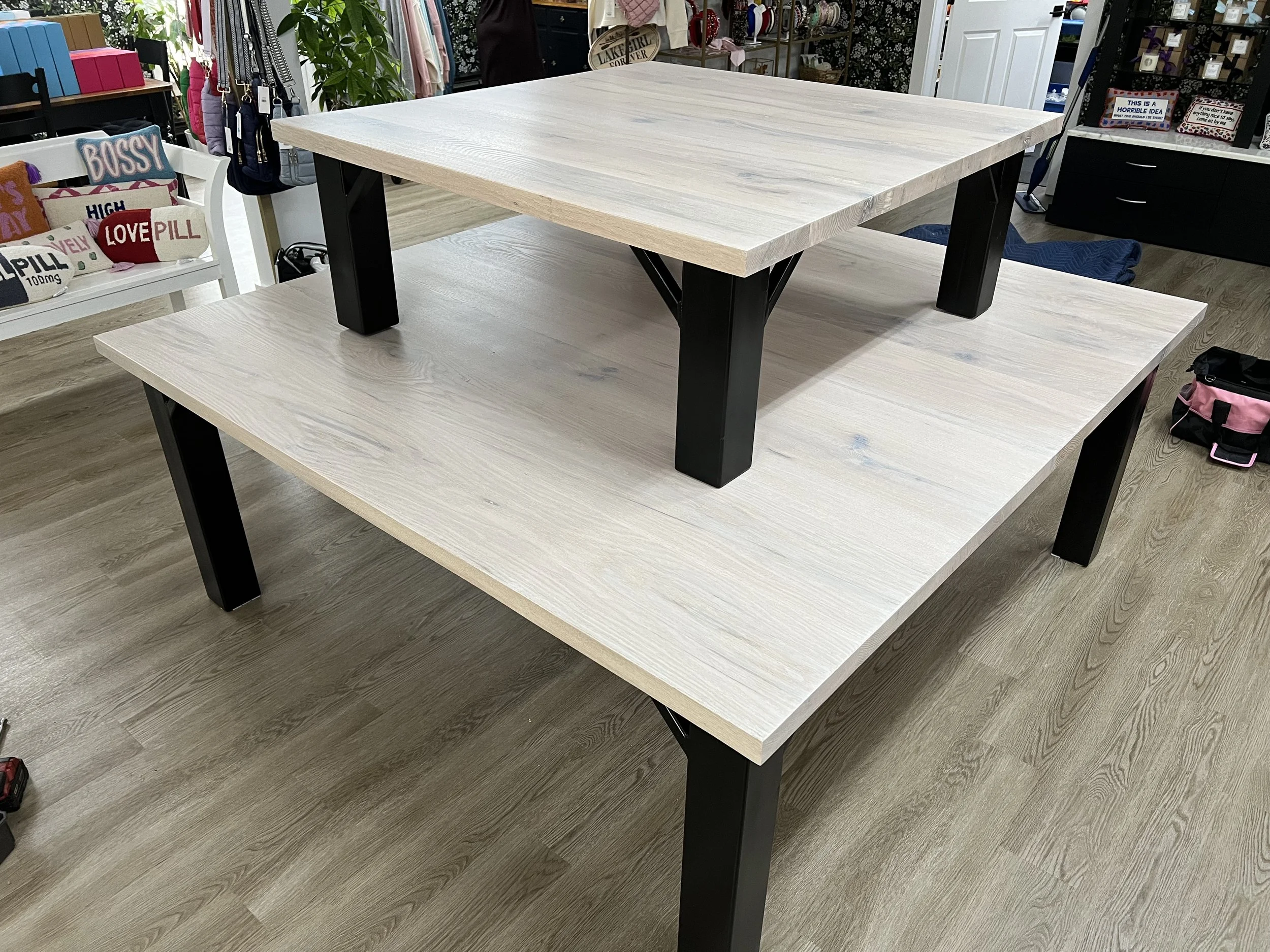 A two-tiered wooden table with black metal legs, placed on a light wood floor in a store with shelves and accessories visible in the background.