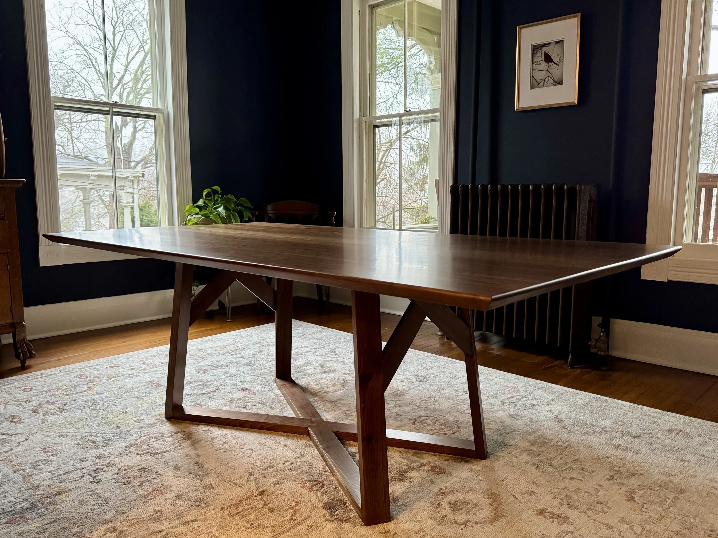 Medium-sized wooden dining table with angled legs placed on a patterned beige area rug in a room with dark blue walls, large windows, and framed artwork, with a potted plant near the window.