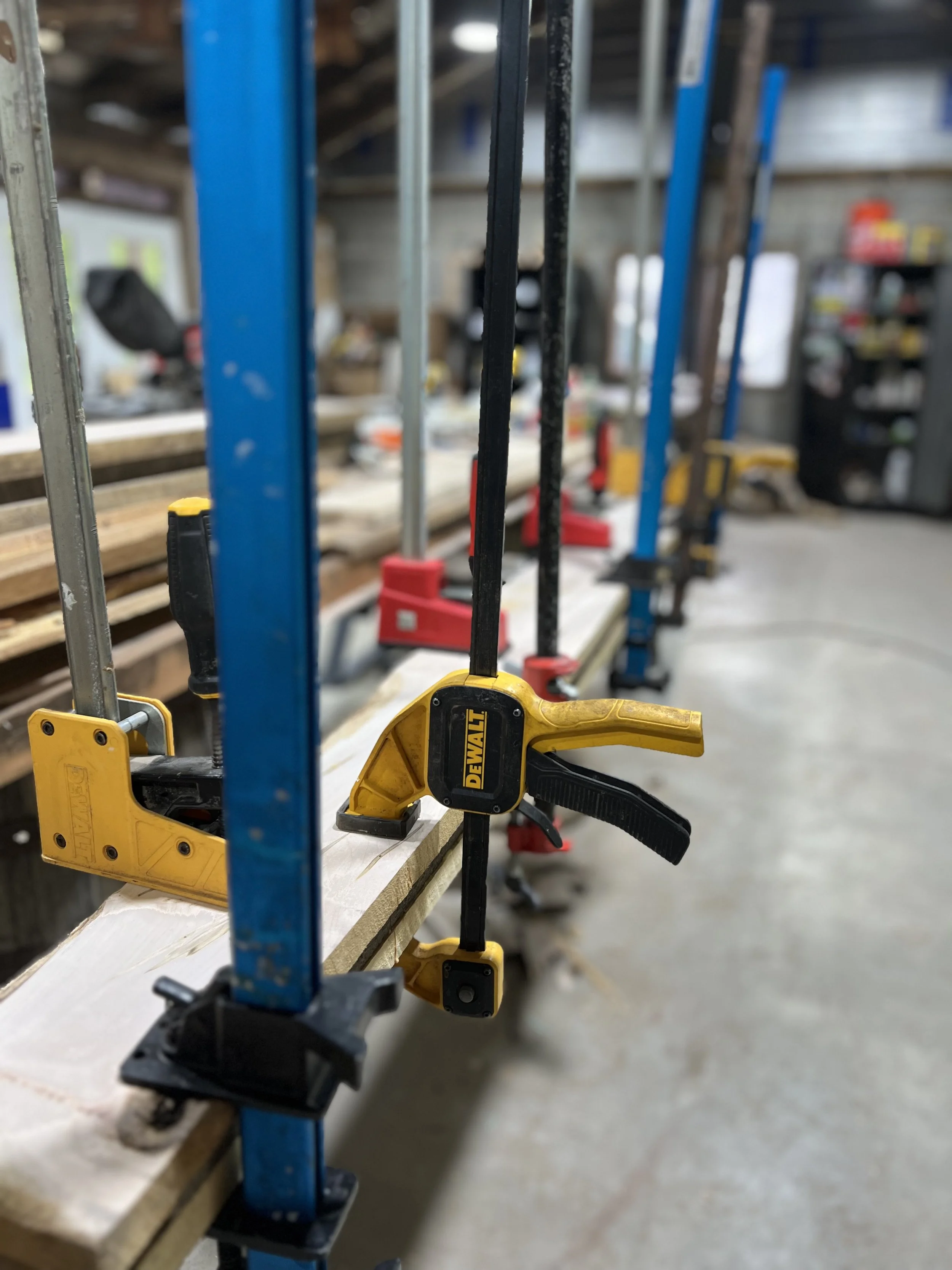 Clamps attached to a long piece of wood on a workbench in a woodworking shop.