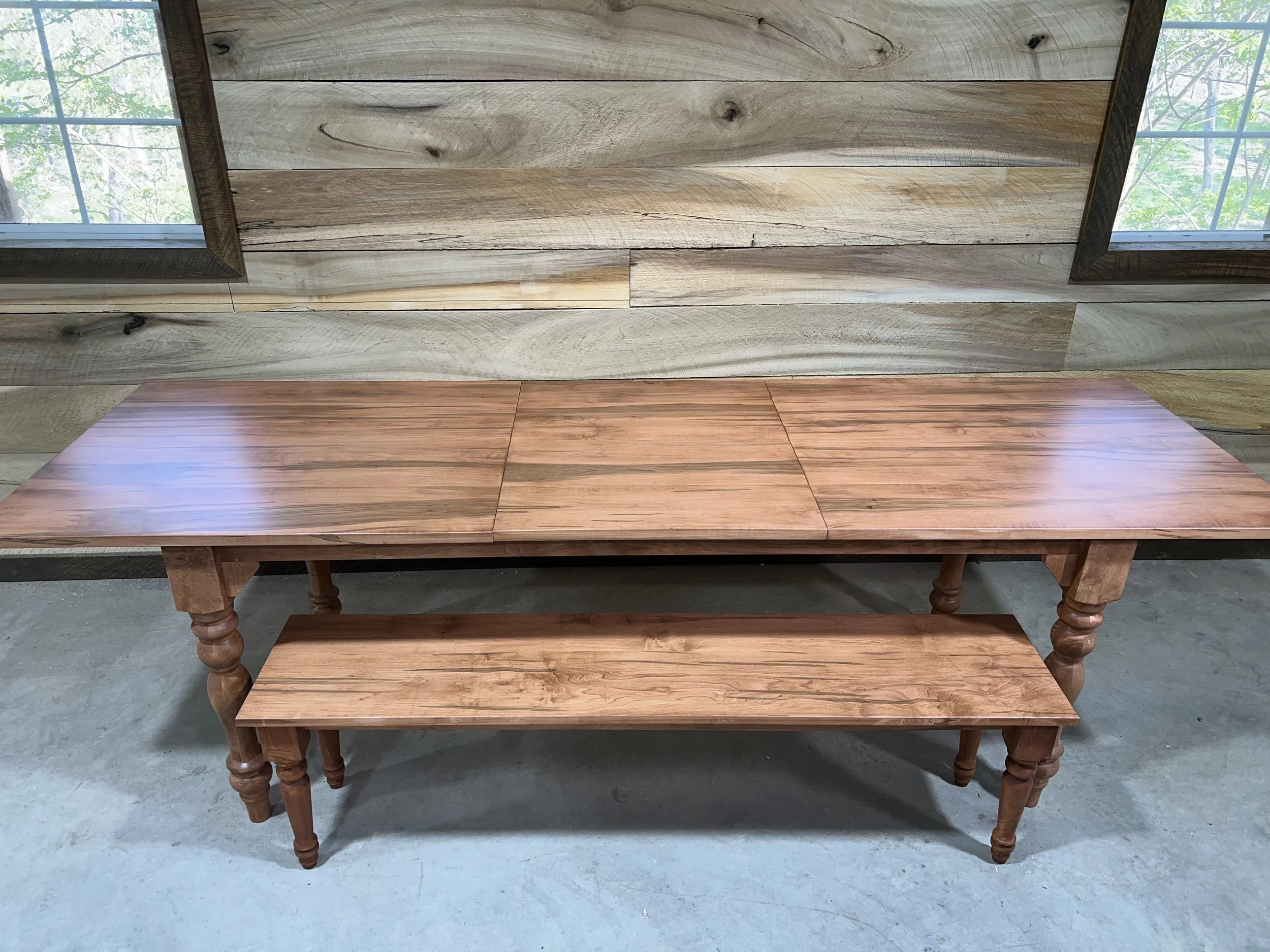 A wooden dining table with a matching bench, positioned in front of a rustic wooden wall with two large windows on either side, allowing natural light to fill the room.