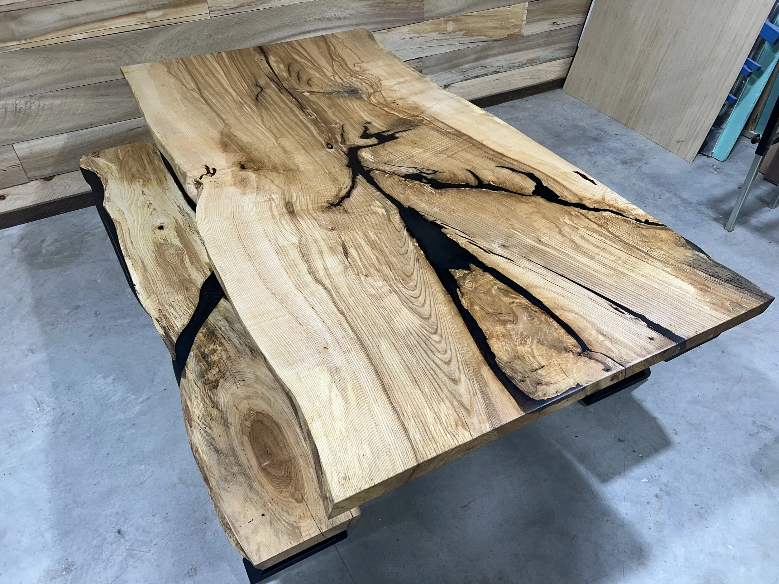 A large, unfinished wooden table with a natural, live edge and black epoxy resin filling the cracks. The table has a polished surface highlighting the grain patterns and knots. It is situated on a concrete floor in a workshop or woodworking studio.