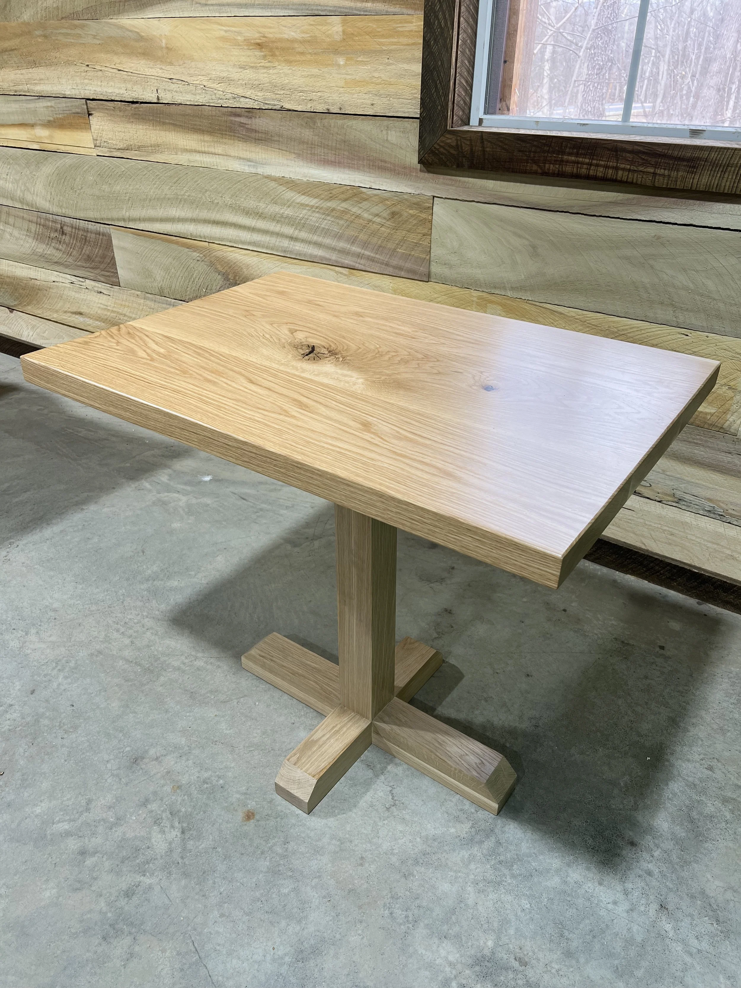 A wooden table with a square top and pedestal base, placed on a concrete floor, in front of a wooden wall with a window.