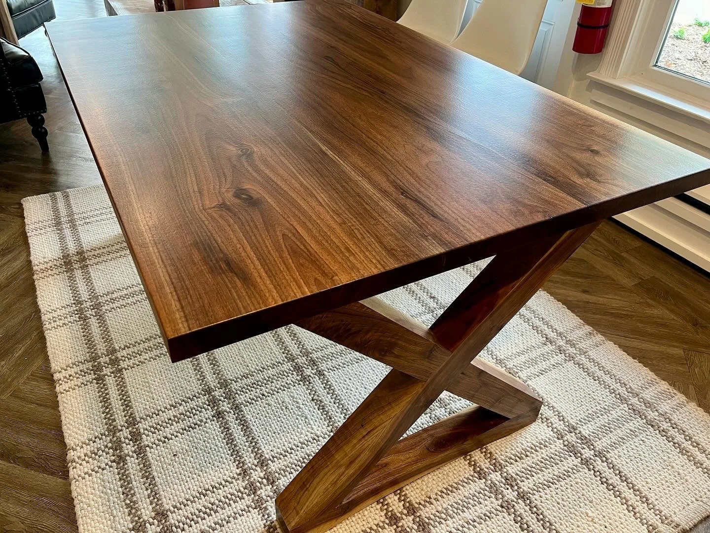 A polished wooden dining table on a cream plaid rug in a room with a window, a fire extinguisher on the wall, and wooden flooring.