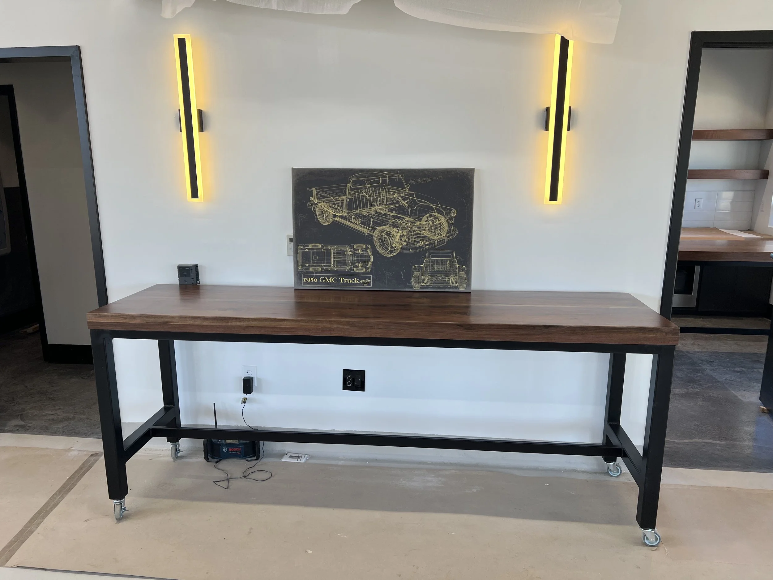 A modern desk with a wooden top and black metal frame, featuring two yellow LED light fixtures on a white wall, with a vintage car blueprint artwork and various electrical outlets and devices.