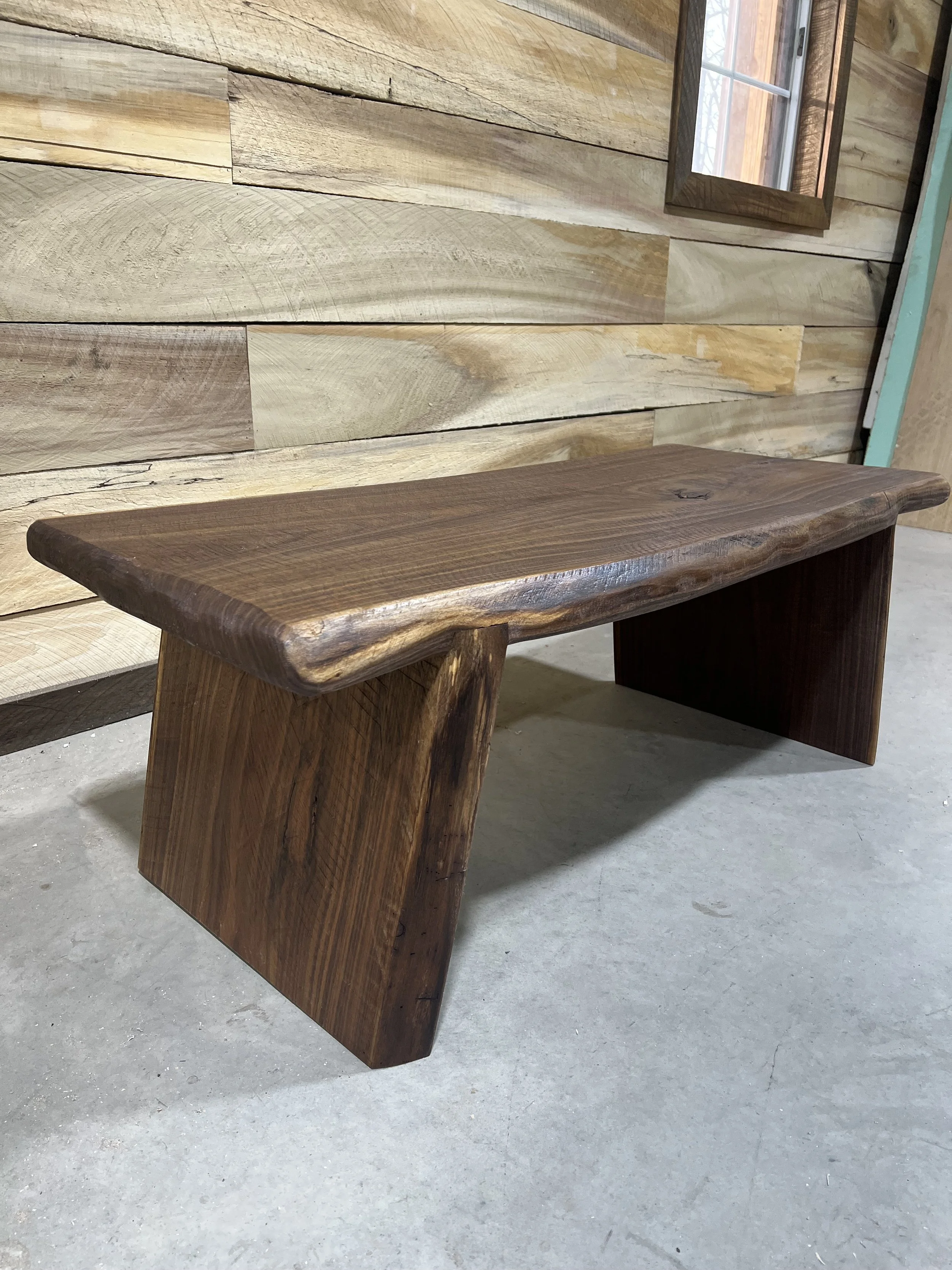 A wooden bench made of dark stained wood, with a smooth, curved top and straight supports, placed on a concrete floor against a wood-paneled wall.
