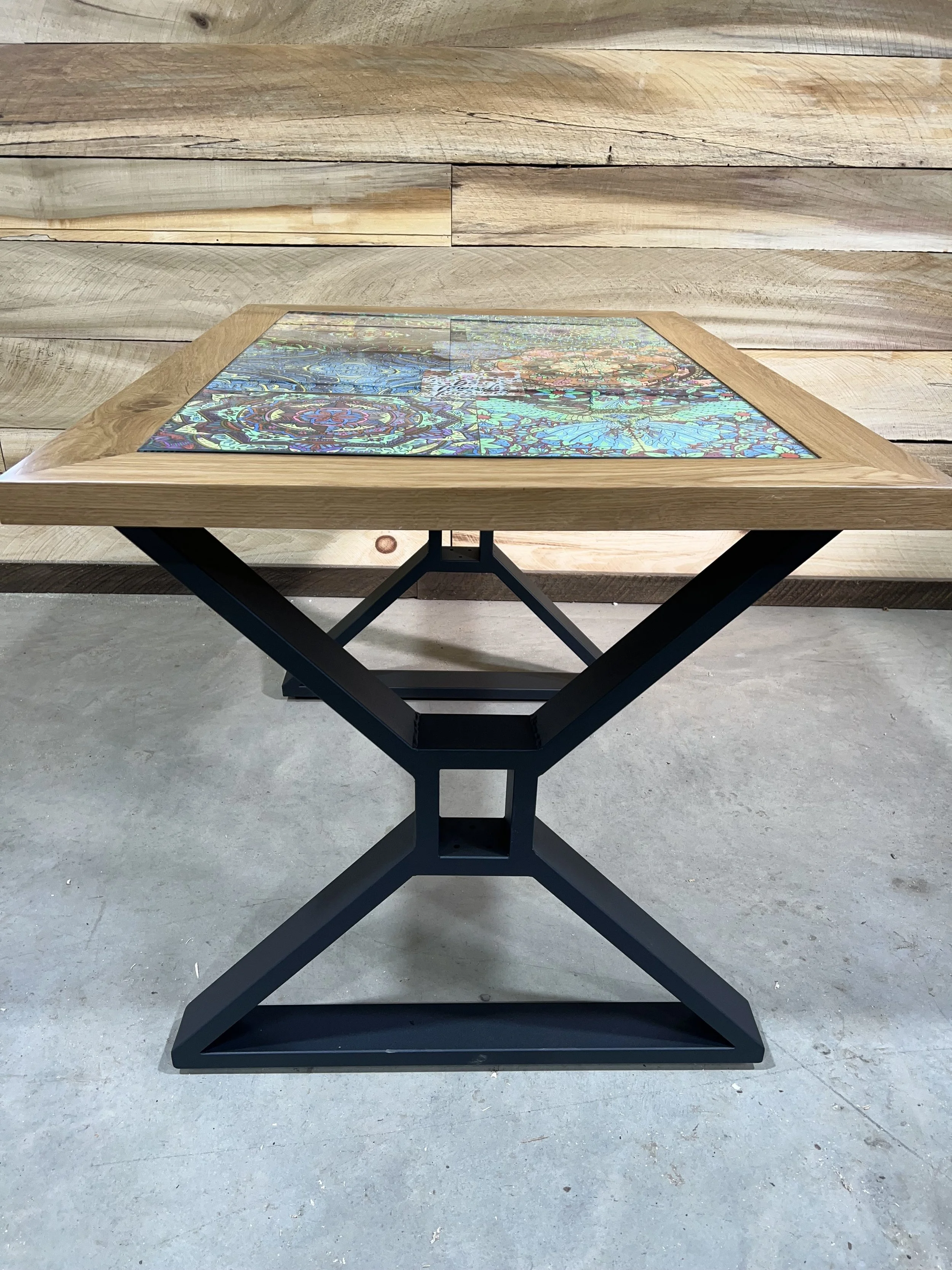 A square table with a black metal base and a wooden border, featuring a glass top with colorful, abstract patterns underneath.