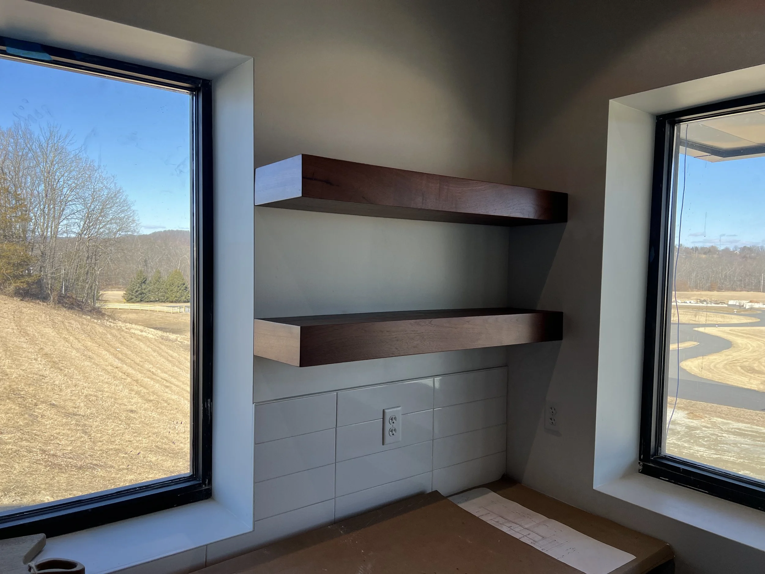 Interior view of a room with two large windows, wooden shelves on a light-colored wall, and an electrical outlet below the shelves.