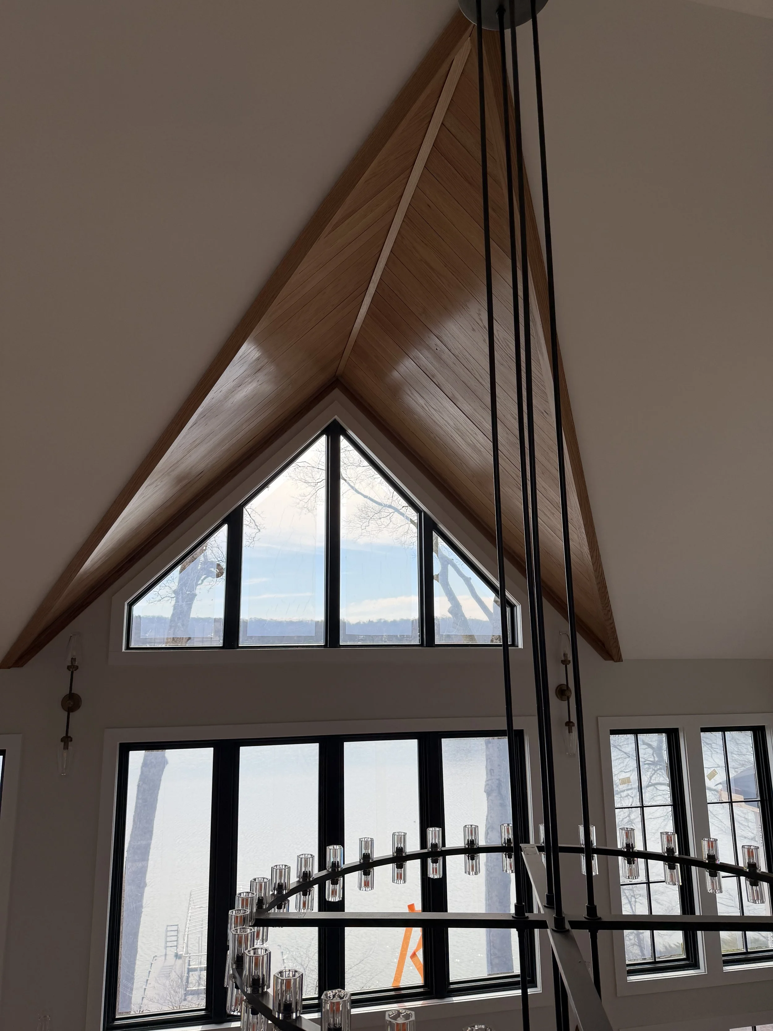 Interior view of a room with large windows and a high, angled wooden ceiling. A modern chandelier made of metal and glass is in the foreground.