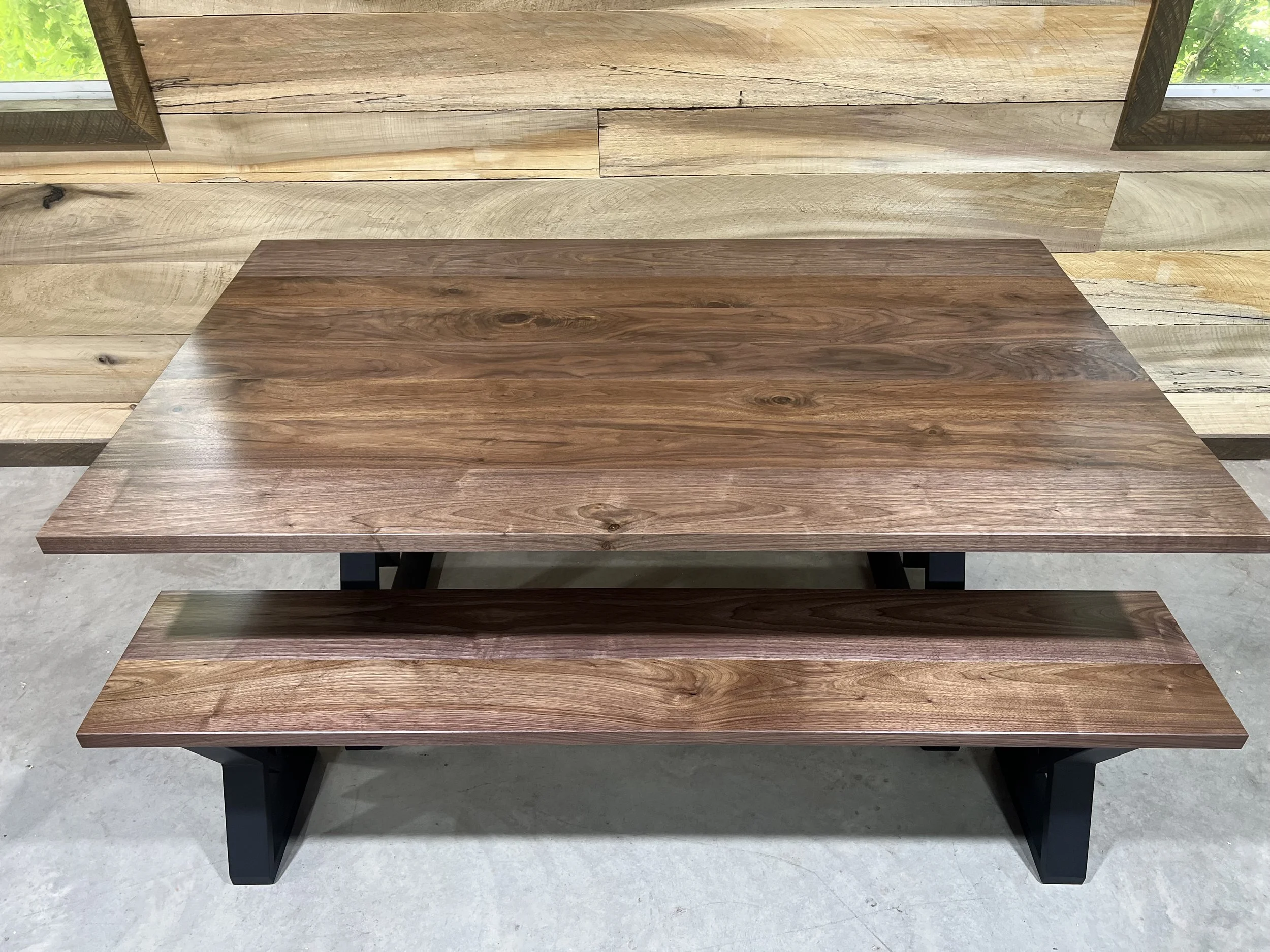 A wooden table with a matching wooden bench, both with natural wood grain finish and dark legs, on a concrete floor with a wood-paneled wall behind them.