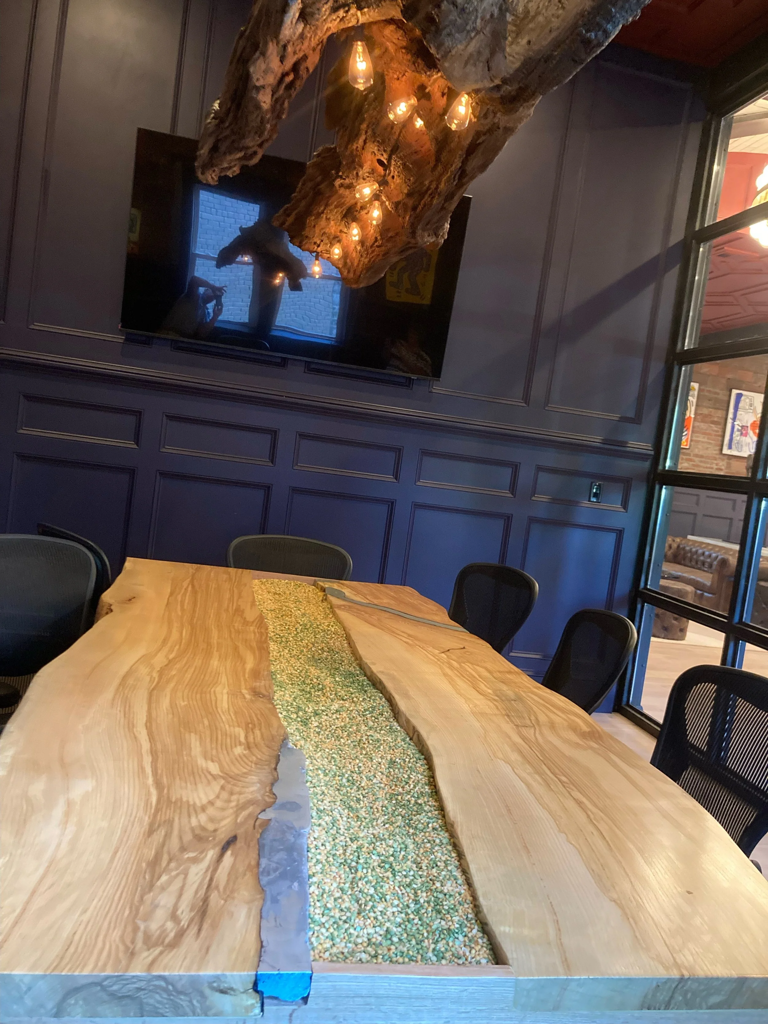 A large wooden conference table with a center filled with multicolored popcorn or tiny beads, in a room with dark blue paneled walls and a mounted flat-screen TV. Above the table hangs a rustic, driftwood light fixture with exposed bulbs, and a windo