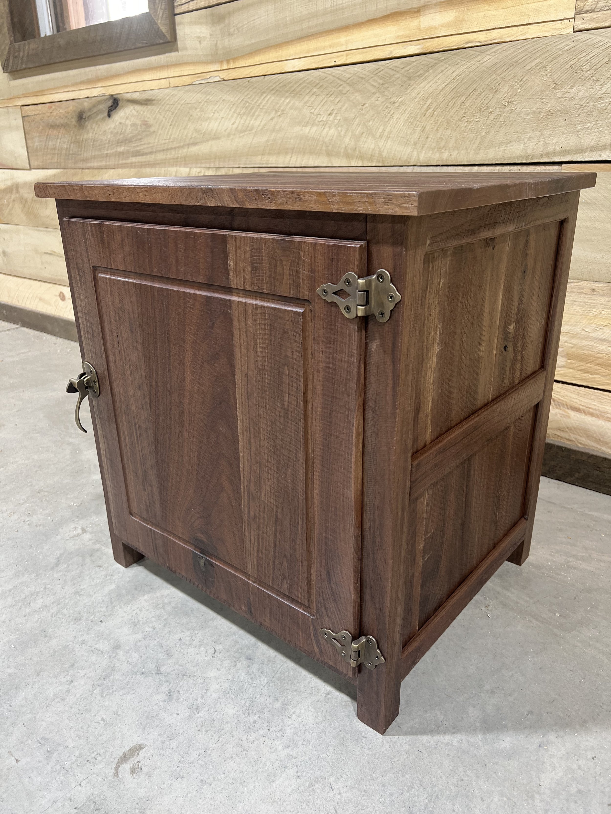 A wooden cabinet standing on a concrete floor, with a natural wood panel wall in the background.