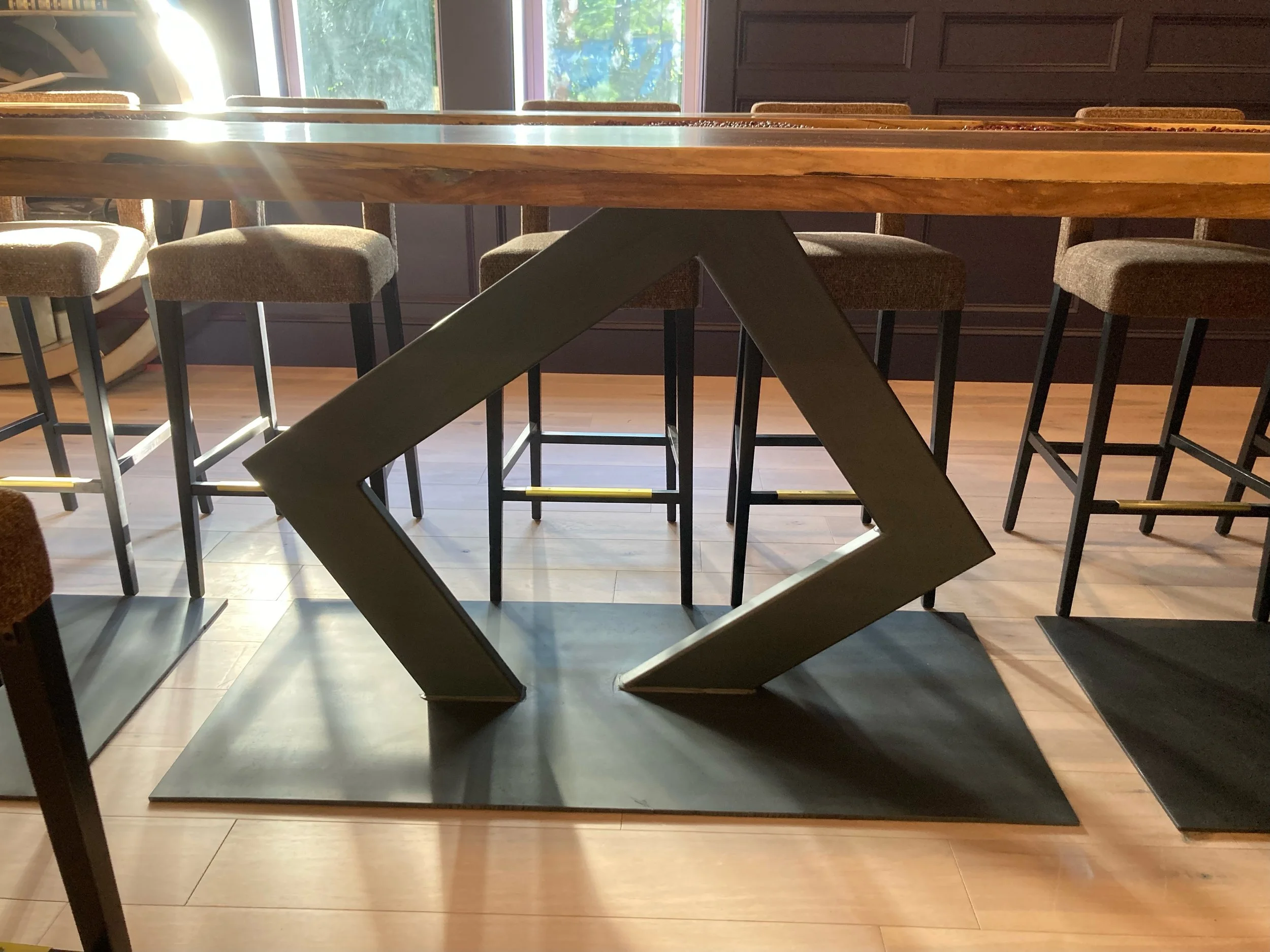 A black metal geometric sculpture in the shape of a stretched diamond on a black base, positioned on a wooden floor near a dining table with chairs.
