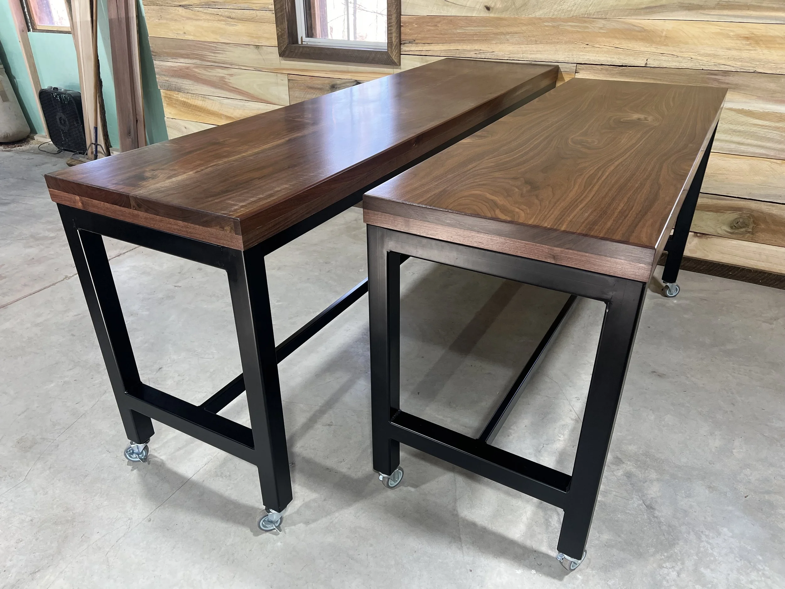 Two wooden tables with black metal frames and caster wheels, placed on a concrete floor in a workshop or showroom.