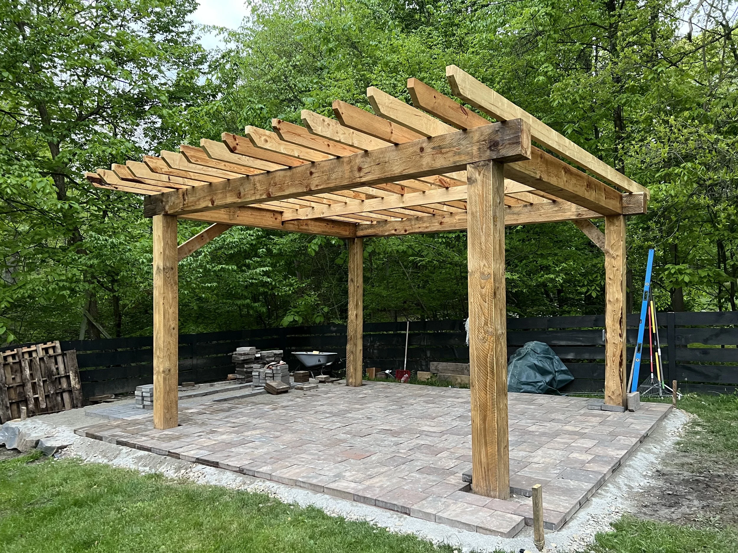 Wooden pergola with a brick patio underneath, surrounded by green trees and a black fence.