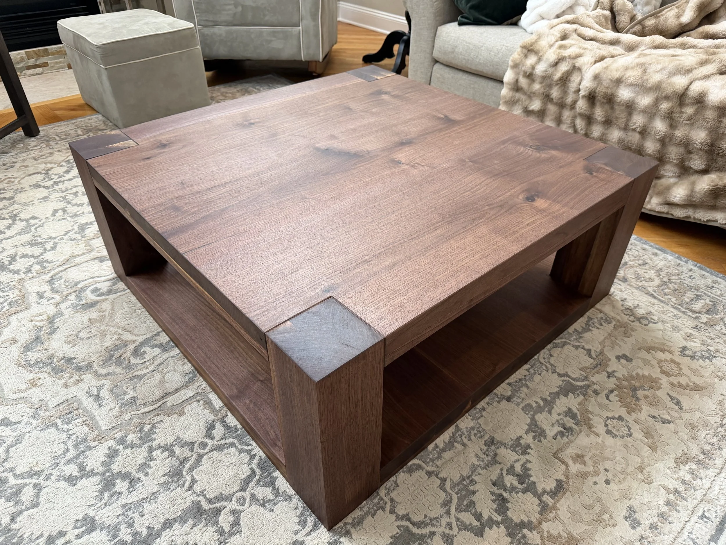 A large square wooden coffee table with open storage space underneath, placed on a patterned rug in a living room.