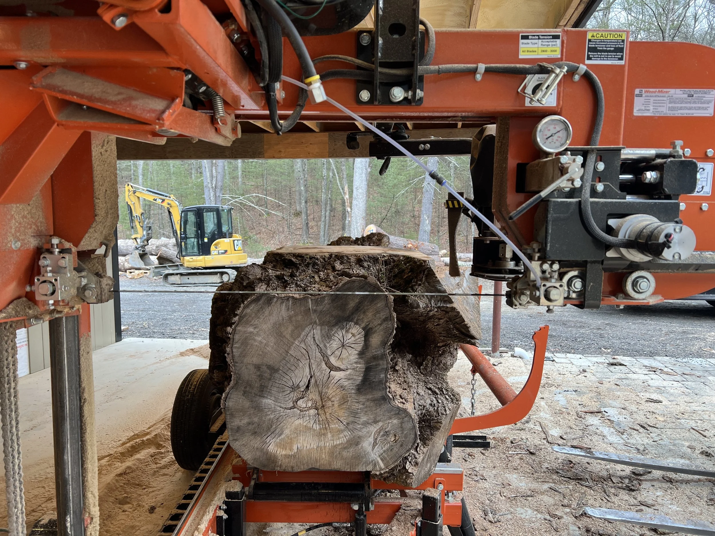 A large band saw cutting through a thick piece of wood outdoors, with construction equipment and trees visible in the background.