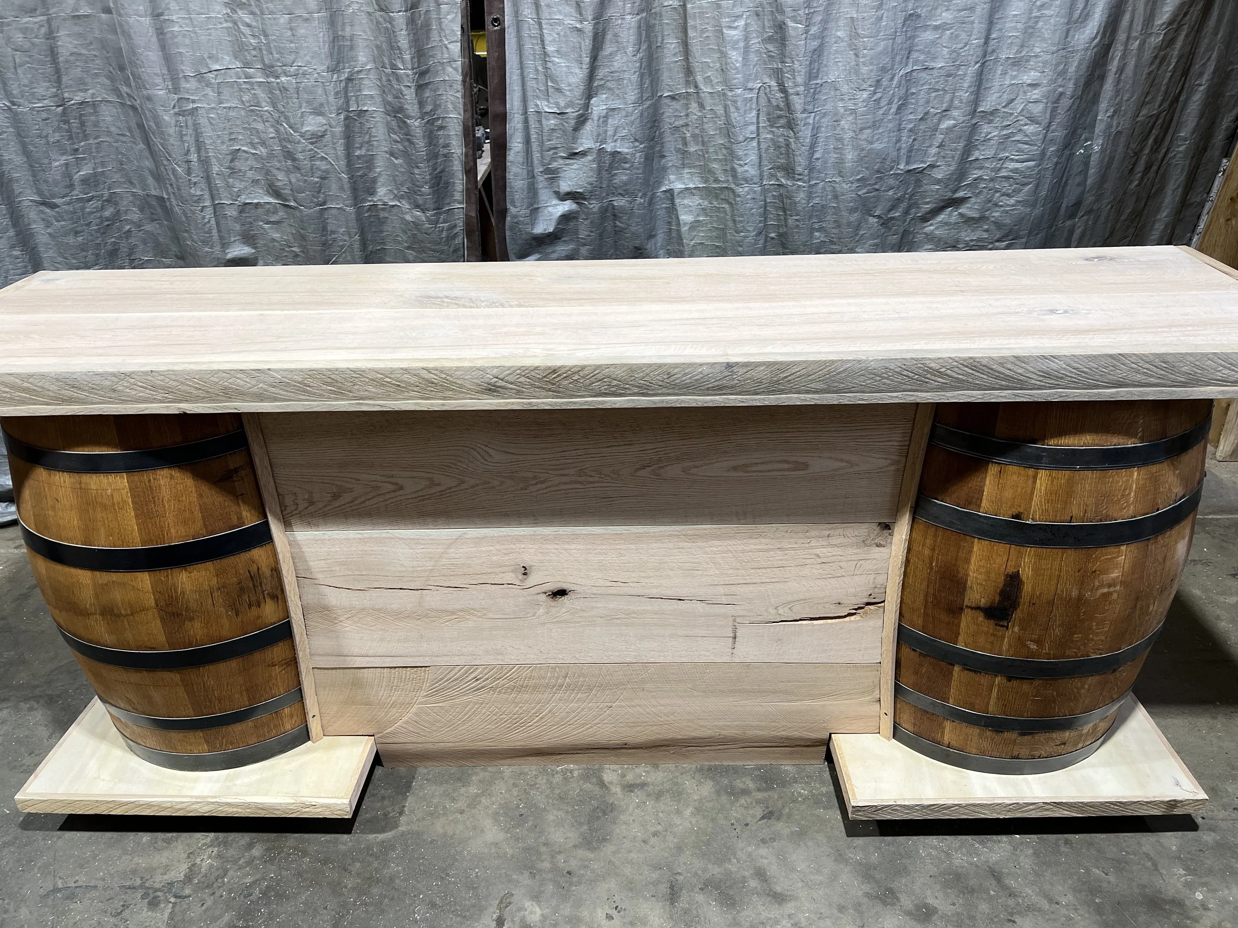A rustic wooden bar or counter with barrel-shaped sides and a light wood top, standing on a concrete floor in front of a gray curtained background.