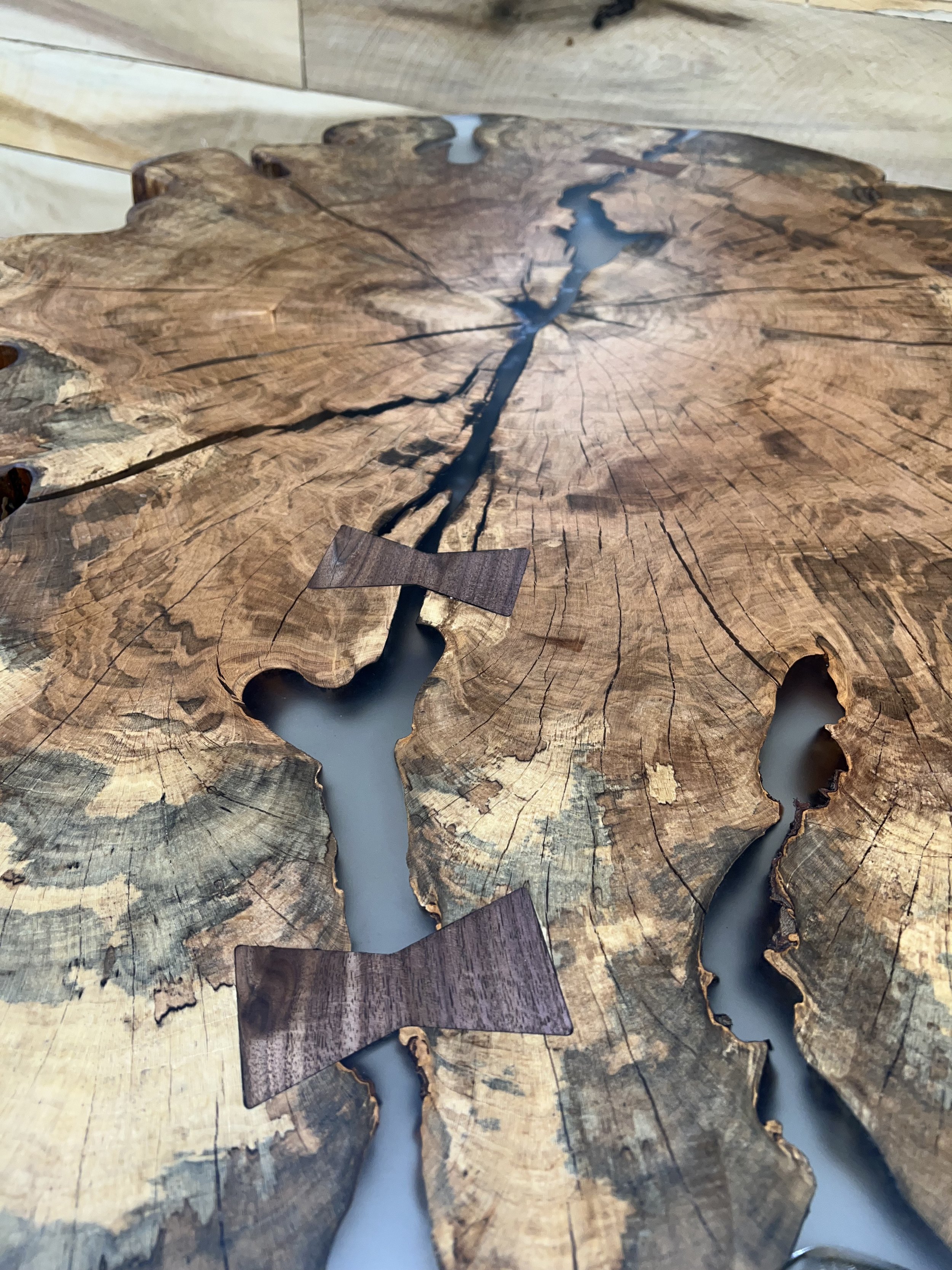 Close-up view of a wooden table made from a large, cross-sectioned tree trunk with visible growth rings and cracks, featuring decorative wooden bow ties and inlay resin highlights.