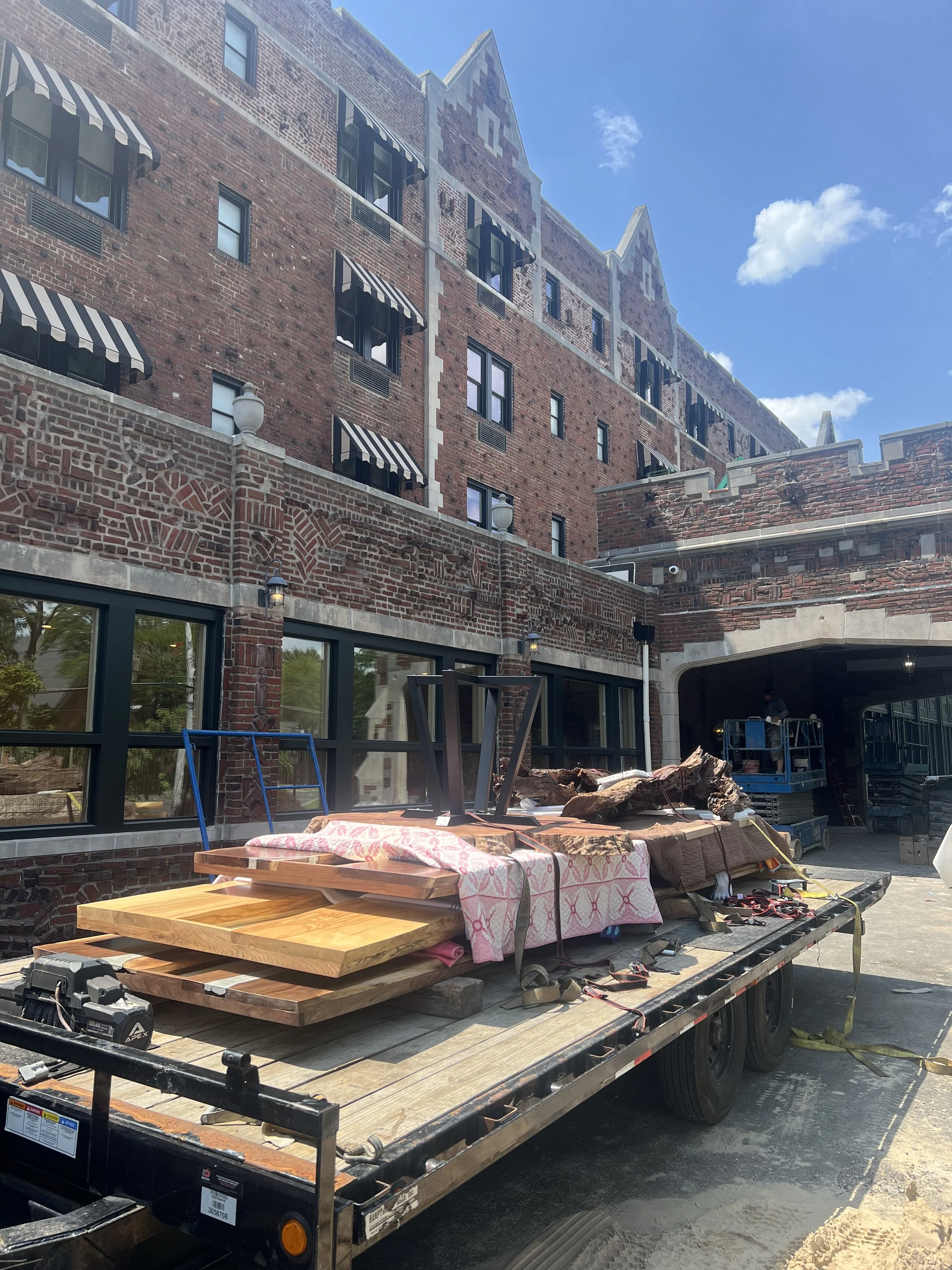 Construction site with a truck loaded with wood and construction materials in front of a large brick building with multiple windows and black and white striped awnings under a blue sky with clouds.