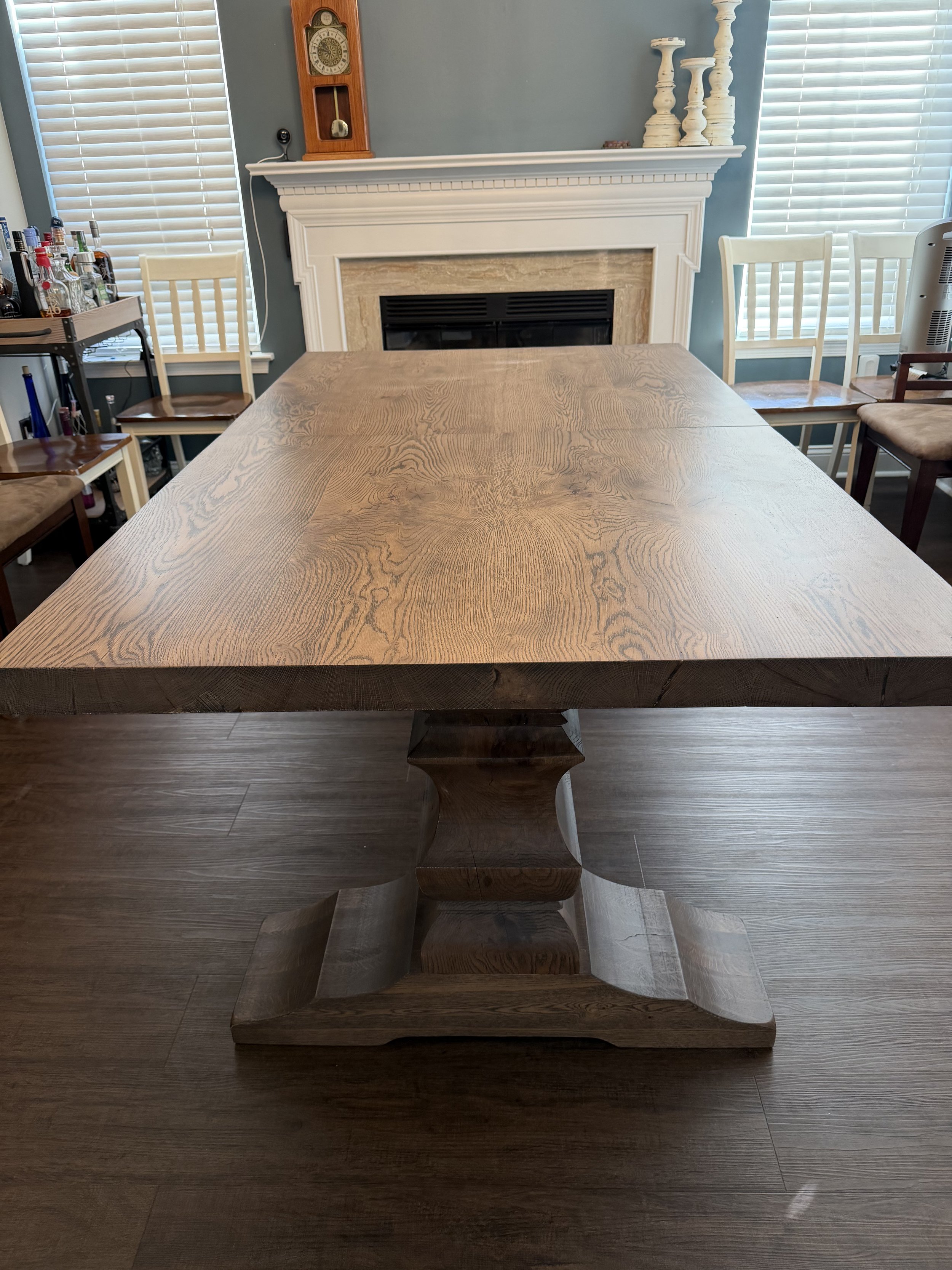 A large wooden dining table in a room with a fireplace, chairs, blinds, and a bar cart.