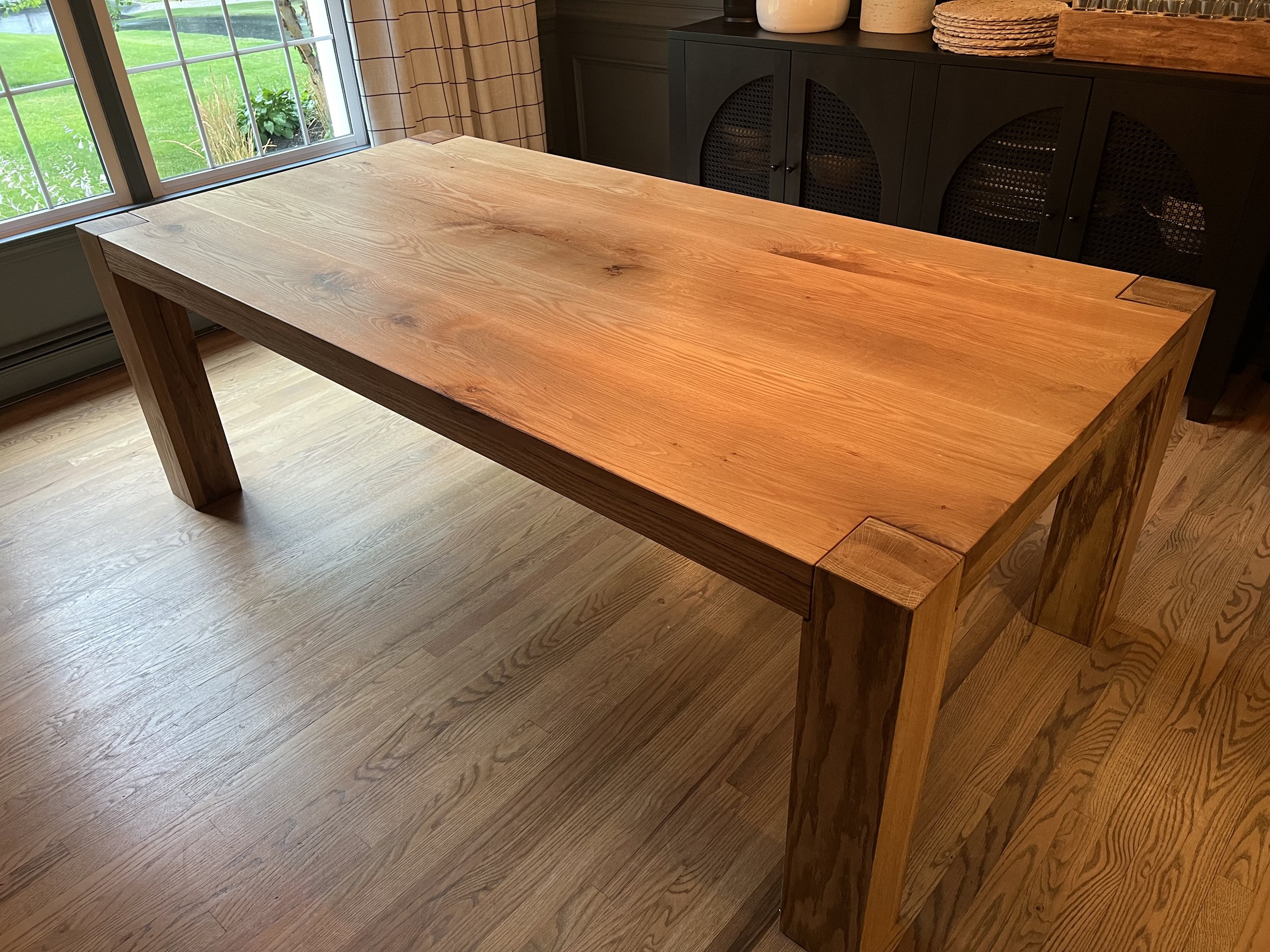 A wooden dining table with a natural finish near a sliding glass door, with a black cabinet and wooden floor in the background.
