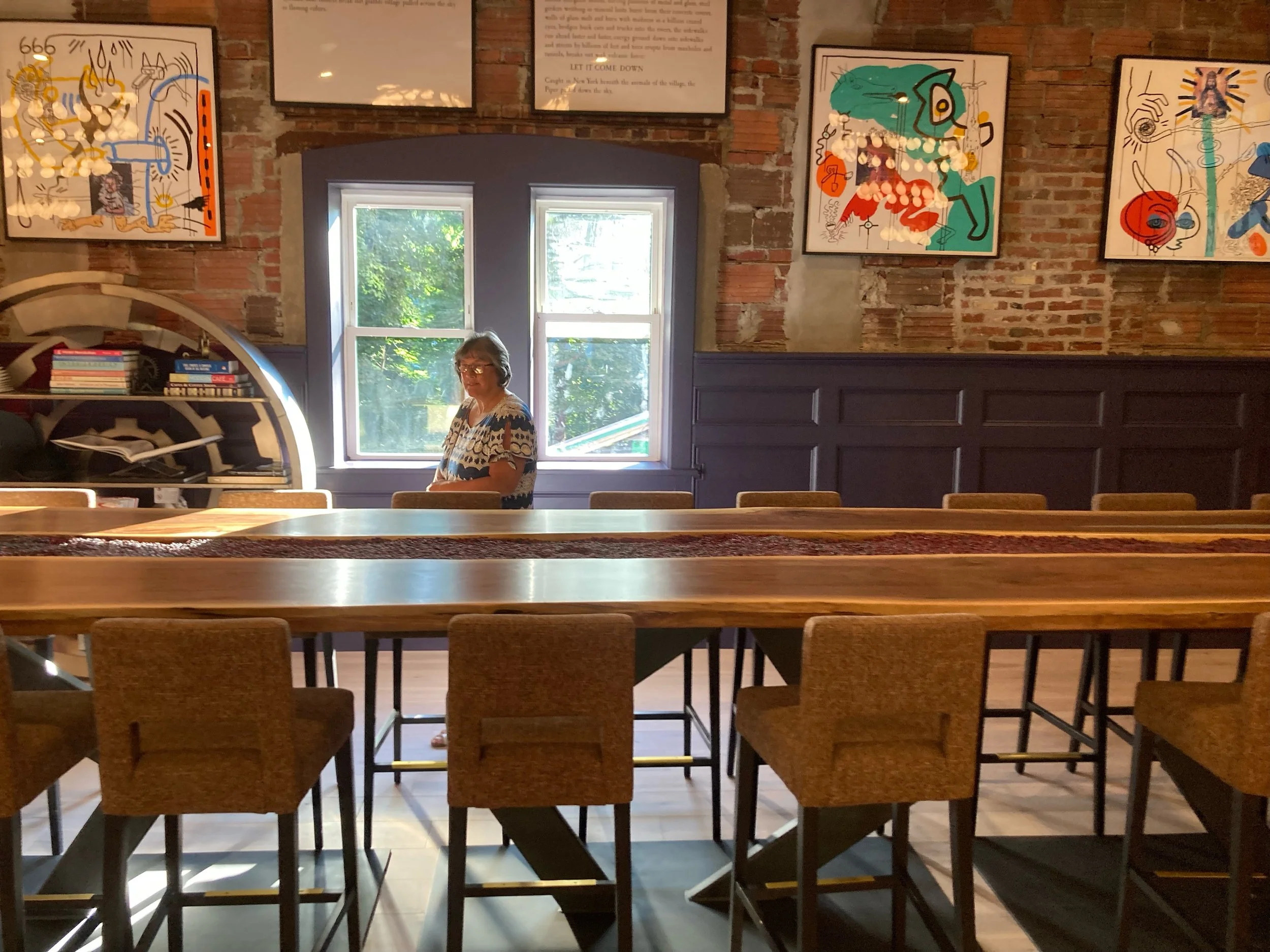 A woman standing inside a room with a large wooden table and chairs, brick walls, colorful abstract artwork, a window with green trees outside, and books on a shelf.